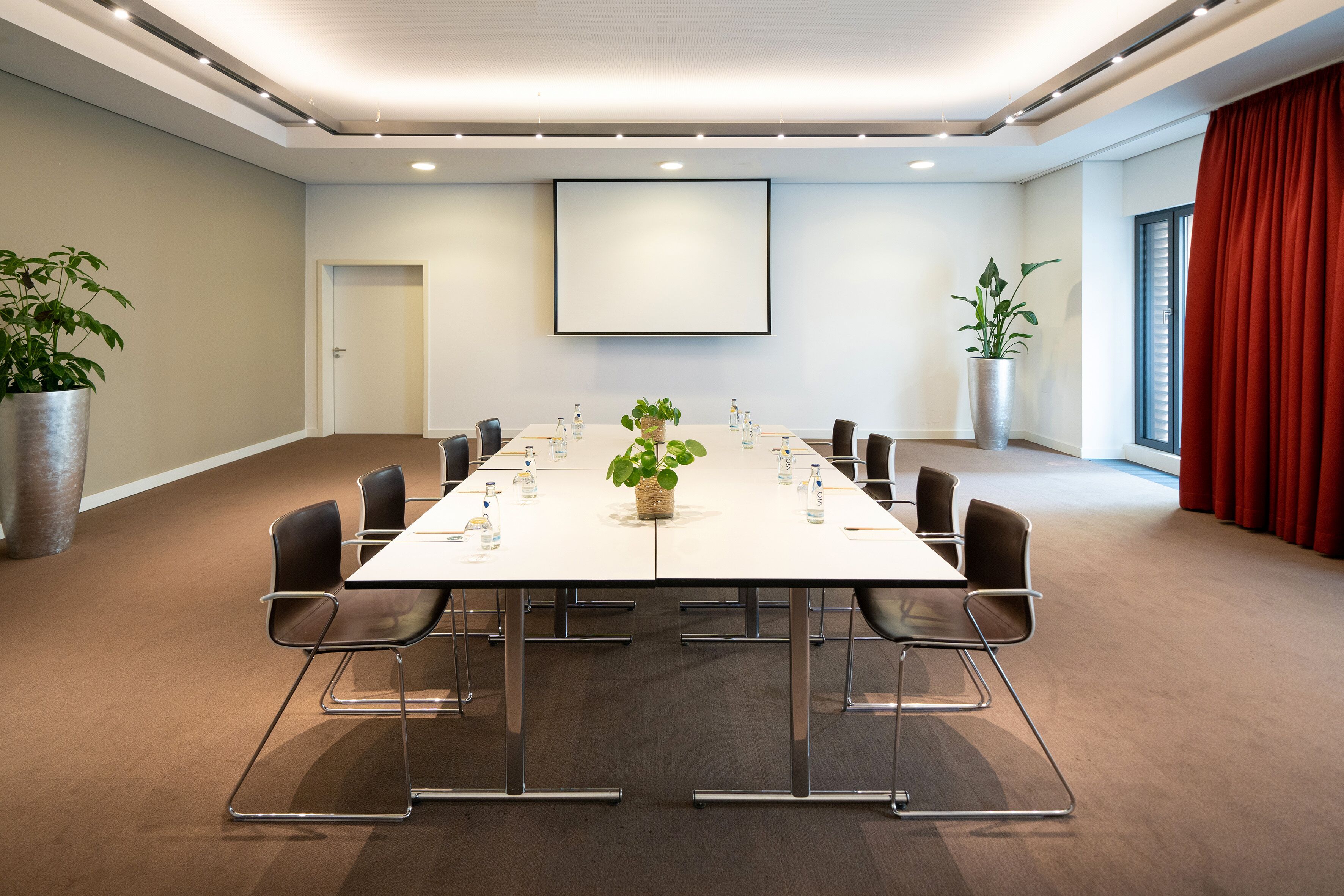 Une salle de conférence avec une longue table, des chaises, des plantes en pot et un grand écran.