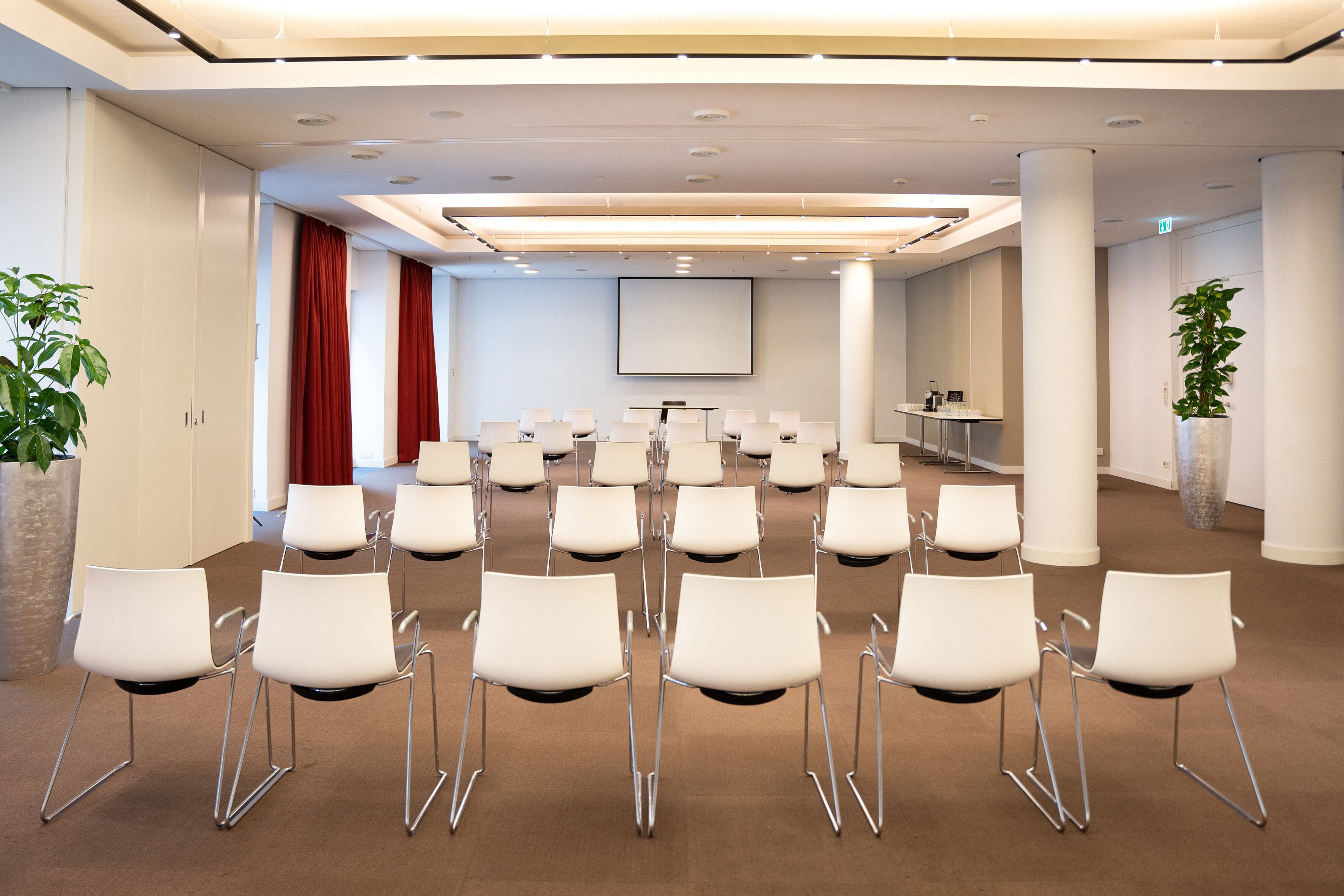 Une salle de conférence avec des chaises blanches disposées en rangées, un grand écran et un pupitre.