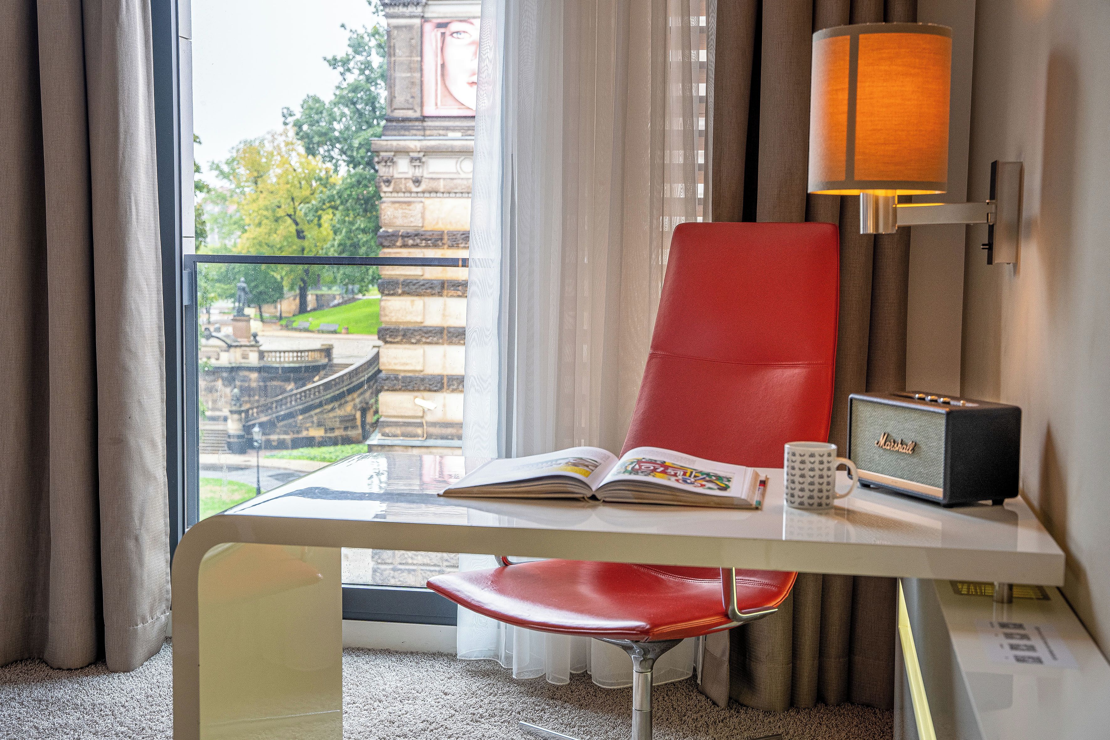 Habitación de hotel con vista a un parque y un edificio, una silla roja, una mesa con un libro y una taza, y un radio Marshall.