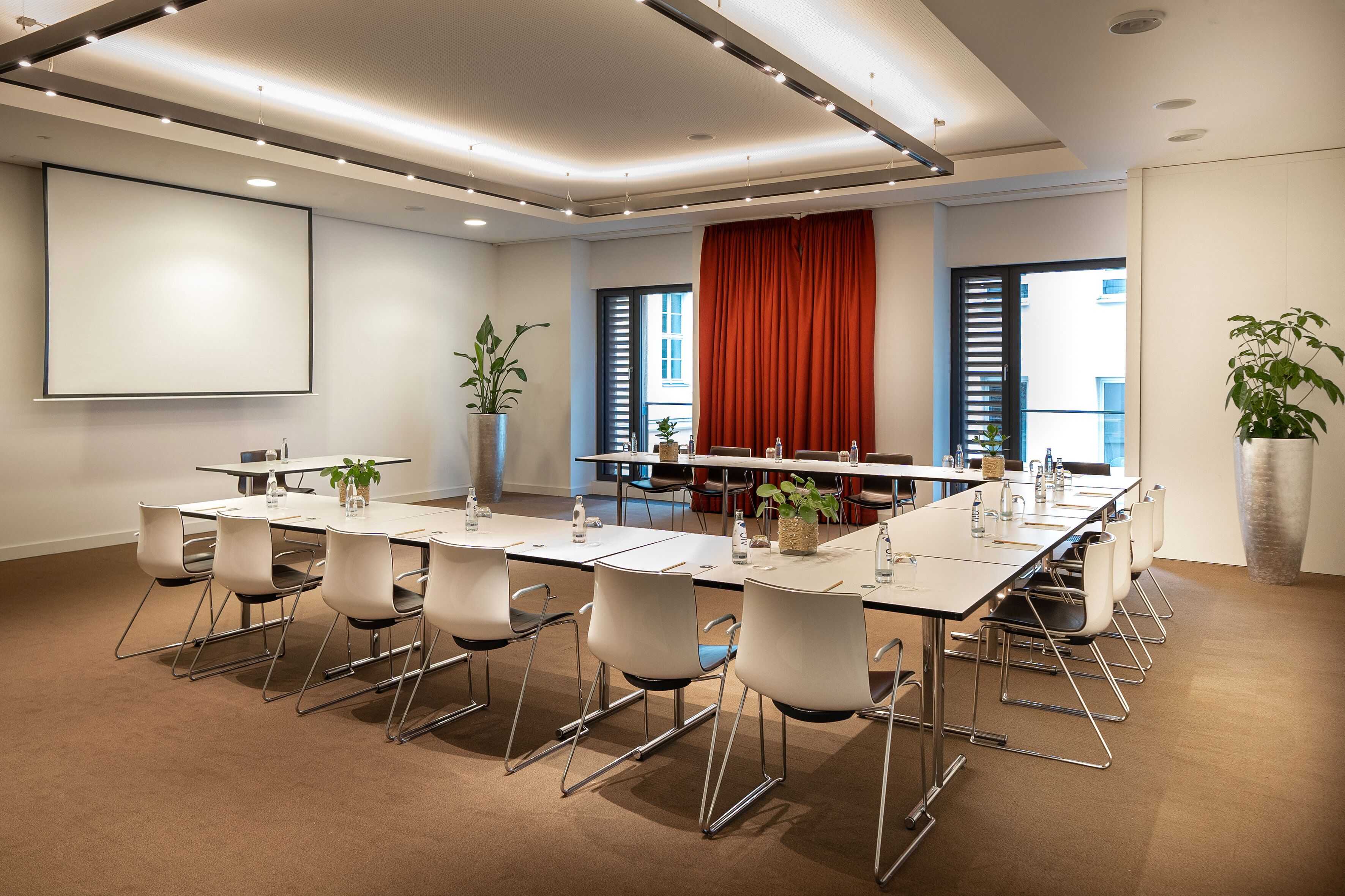 Una sala conferenze con un lungo tavolo, sedie, uno schermo per proiezioni e piante in vaso.