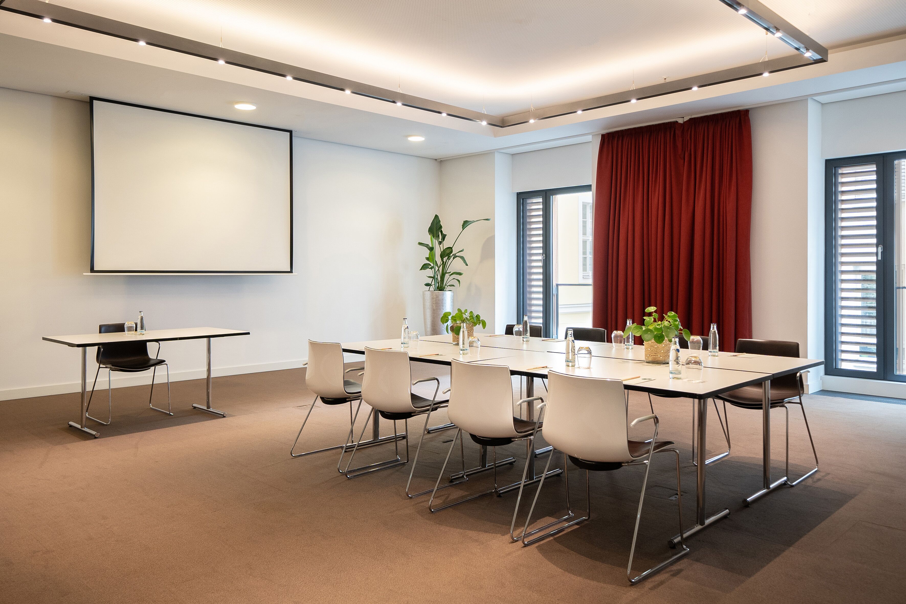 Une salle de conférence équipée d’une grande table, de chaises, d’un écran de projection et d’une plante en pot.