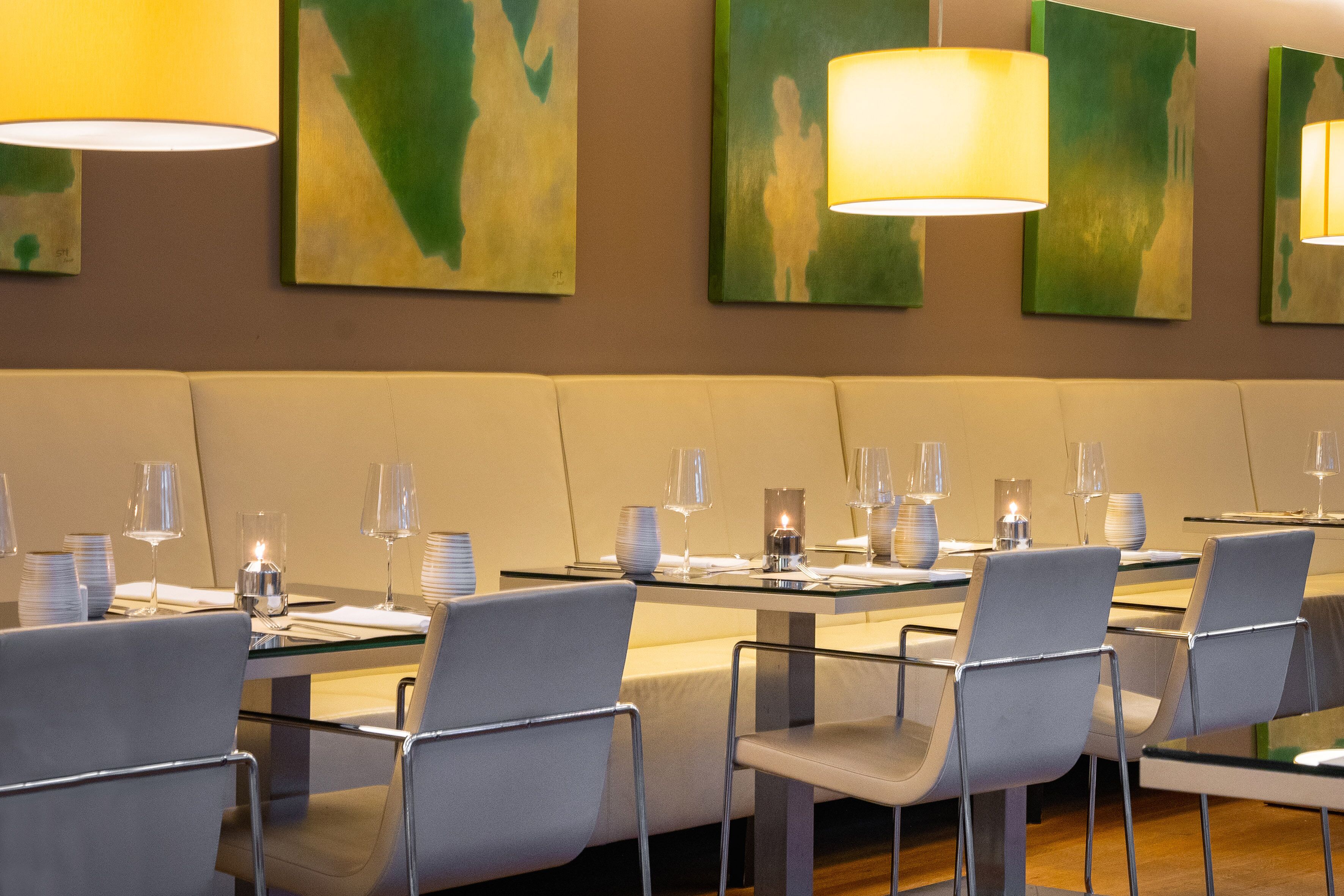 A restaurant with a modern interior, featuring glass tables and metal chairs, with artwork on the walls and pendant lighting.