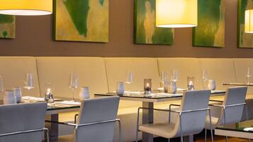 A restaurant with a modern interior, featuring glass tables and metal chairs, with artwork on the walls and pendant lighting.