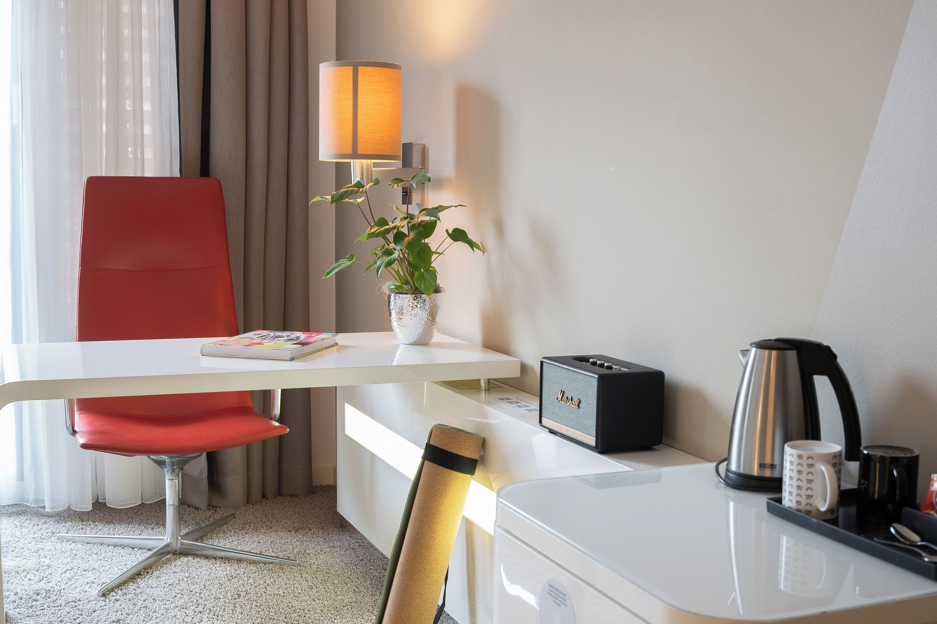 Ein Hotelzimmer mit weißem Schreibtisch, einem roten Sessel, einer Lampe, einer Pflanze, einem Radio, einem Wasserkocher und einem Tablett mit Tasse und Löffel.