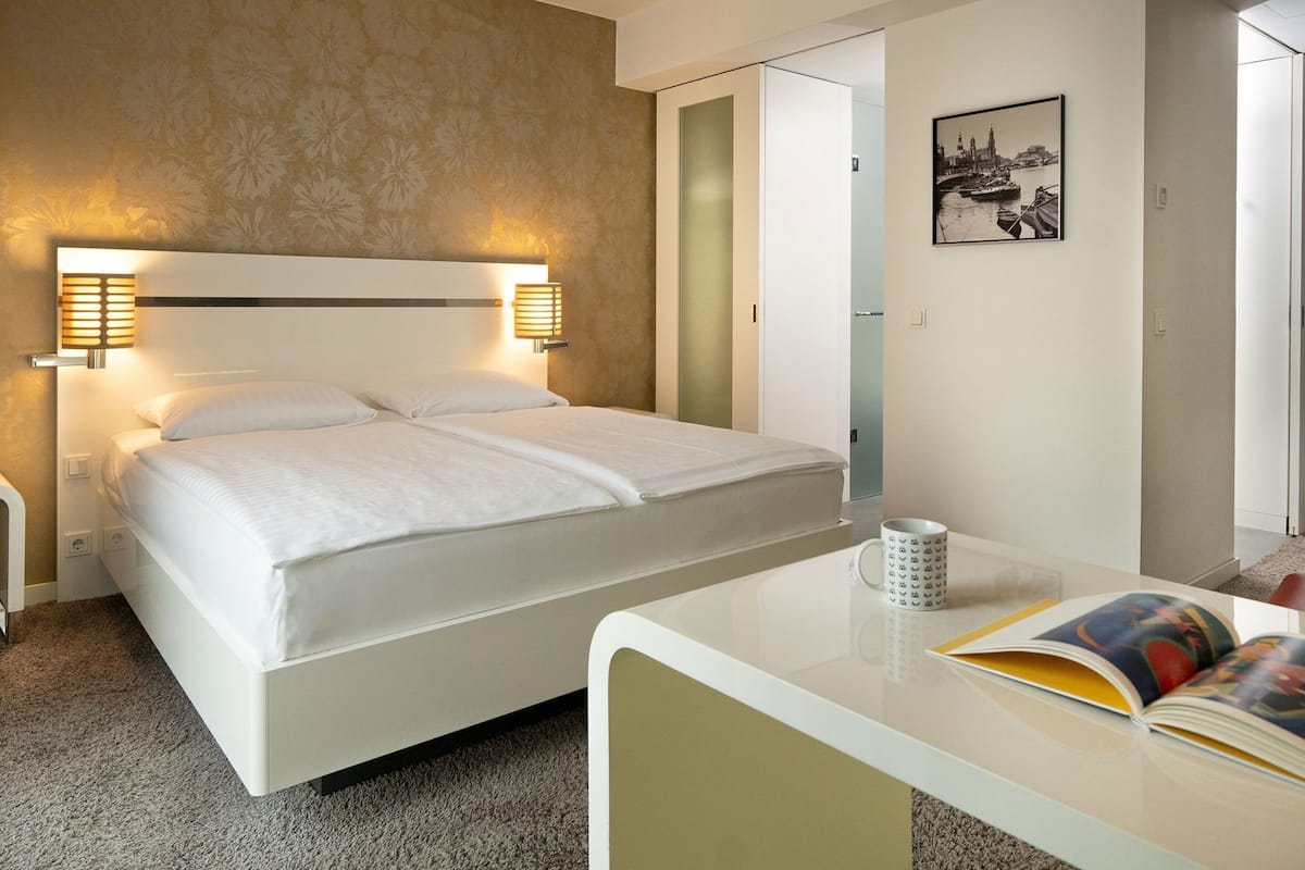 Habitación de hotel con cama, lámparas de noche, un escritorio con una taza y un cuadro en la pared.