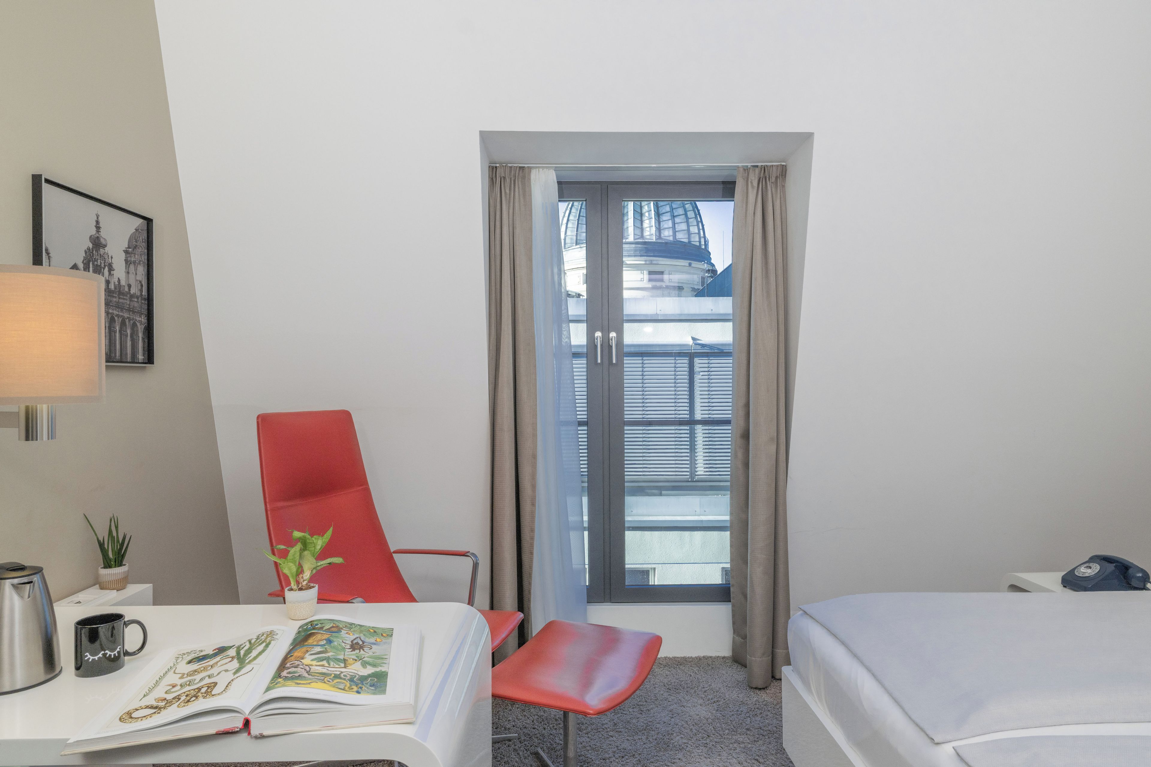 Quarto de hotel com uma cama, uma escrivaninha com luminária, uma cadeira, uma mesa pequena com uma planta e uma janela com cortinas.