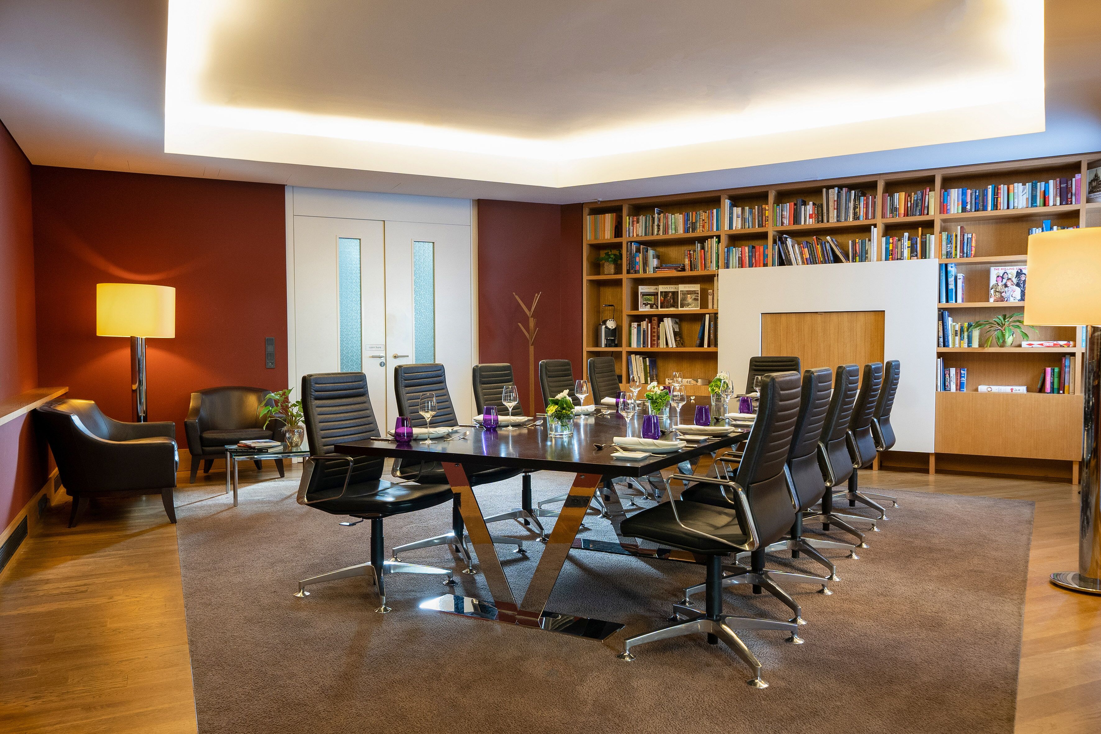 Een vergaderruimte met een lange tafel, stoelen en een boekenplank vol boeken.