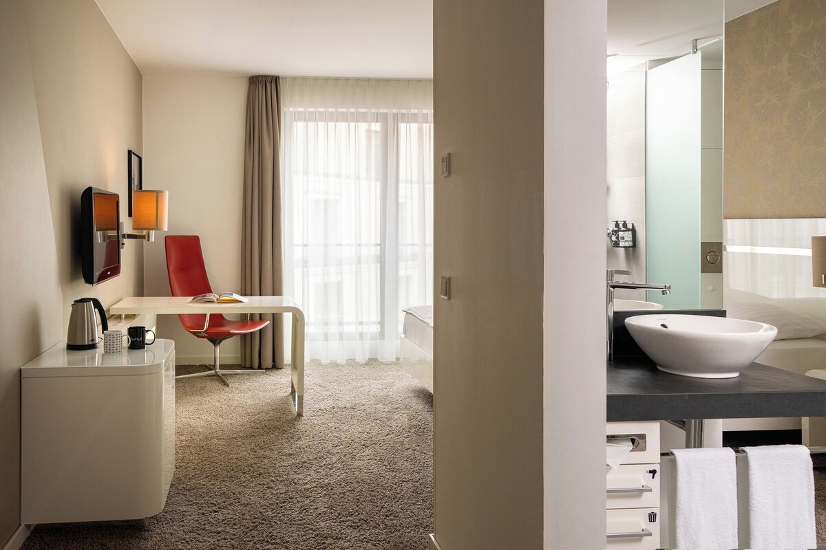 Hotelkamer met bureau, stoel en een moderne badkamer met een groot wastafel en spiegel.