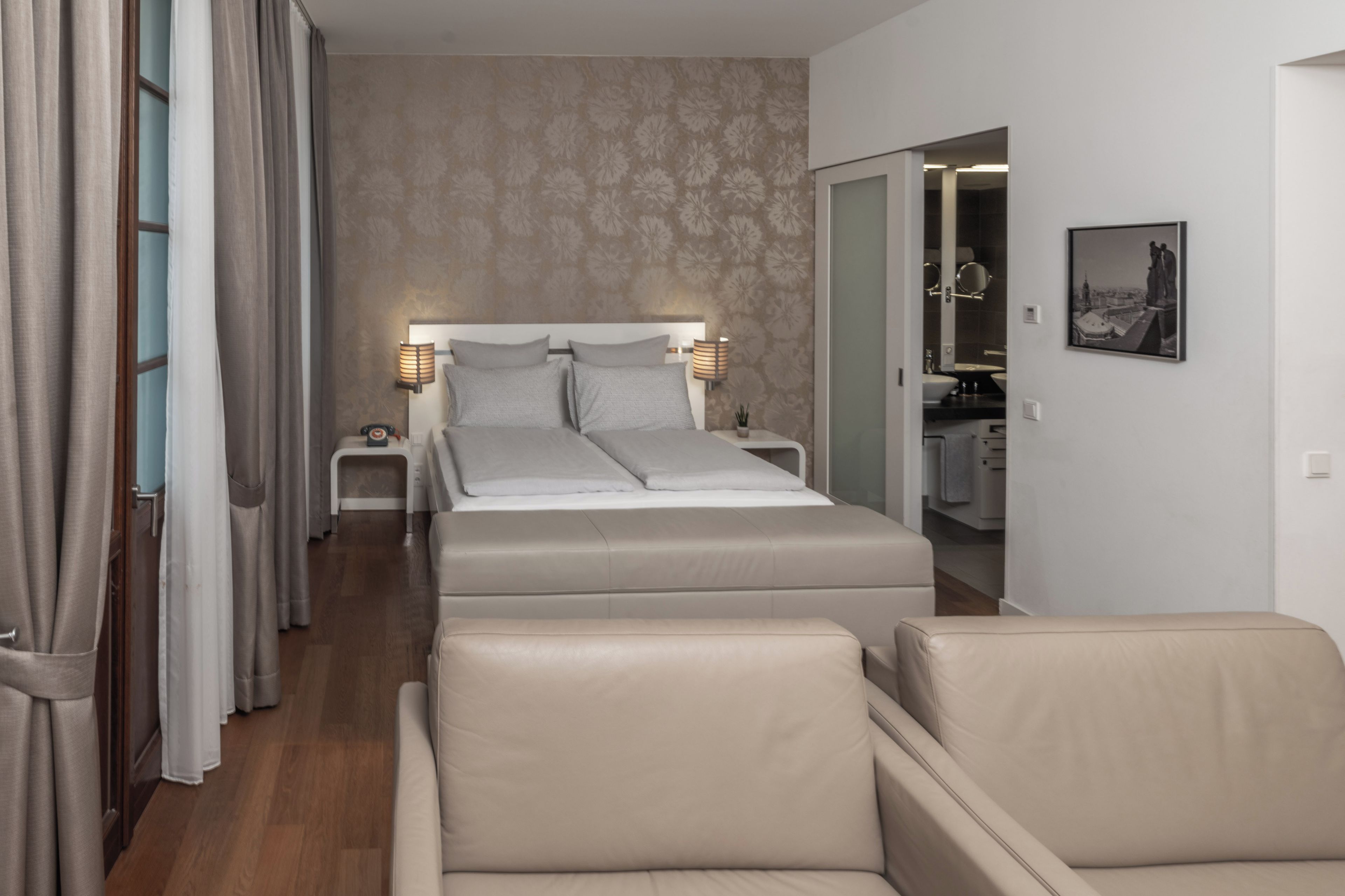 Ein Hotelzimmer mit Bett, Nachttischlampen, einem Nachttisch, einem Sessel und einem Badezimmer mit Spiegel. An der Wand hängt ein gerahmtes Bild.