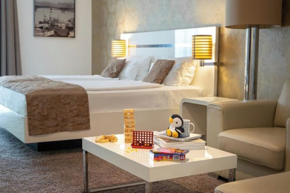 Een hotelkamer met een bed, nachtlampjes, een bijzettafeltje met boeken en een speeltje pinguïn, en een stoel.