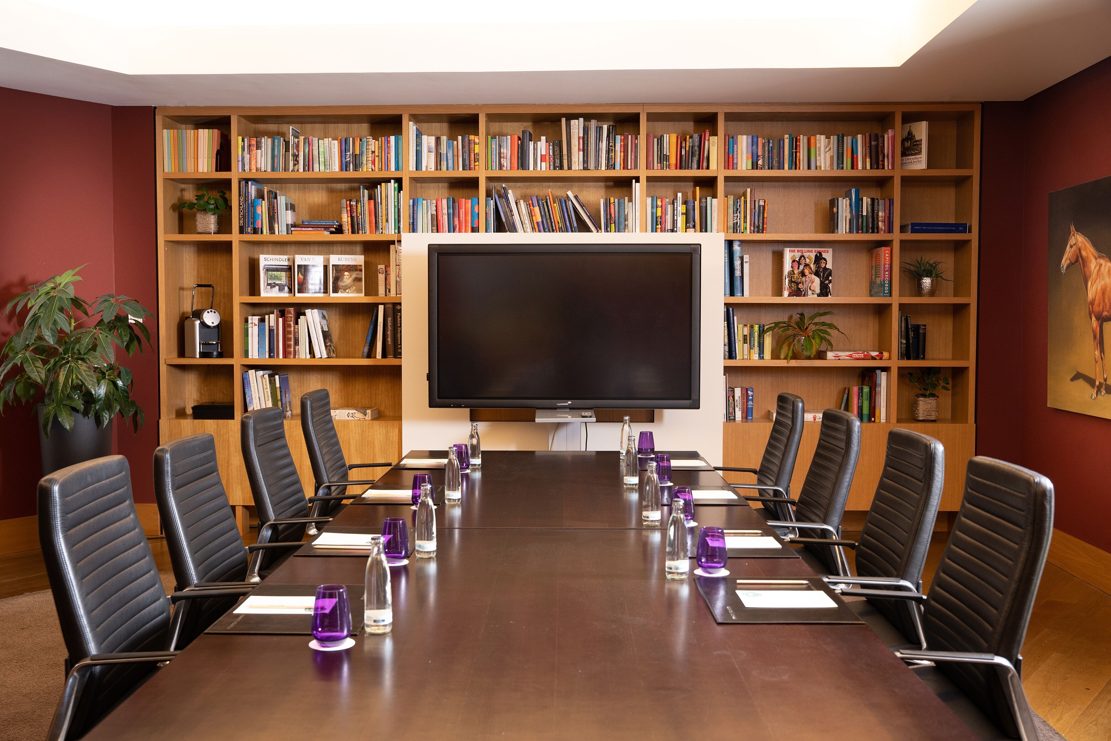 A conference room with a long table, black chairs, a large bookshelf, and a flat-screen TV.