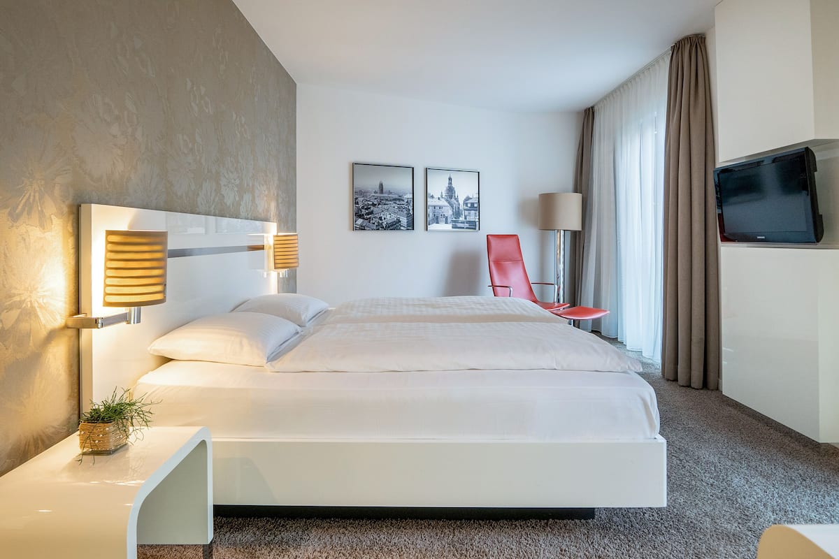 Habitación de hotel moderna con una cama grande, una silla roja, un televisor de pantalla plana y dos cuadros con vistas urbanas en la pared.