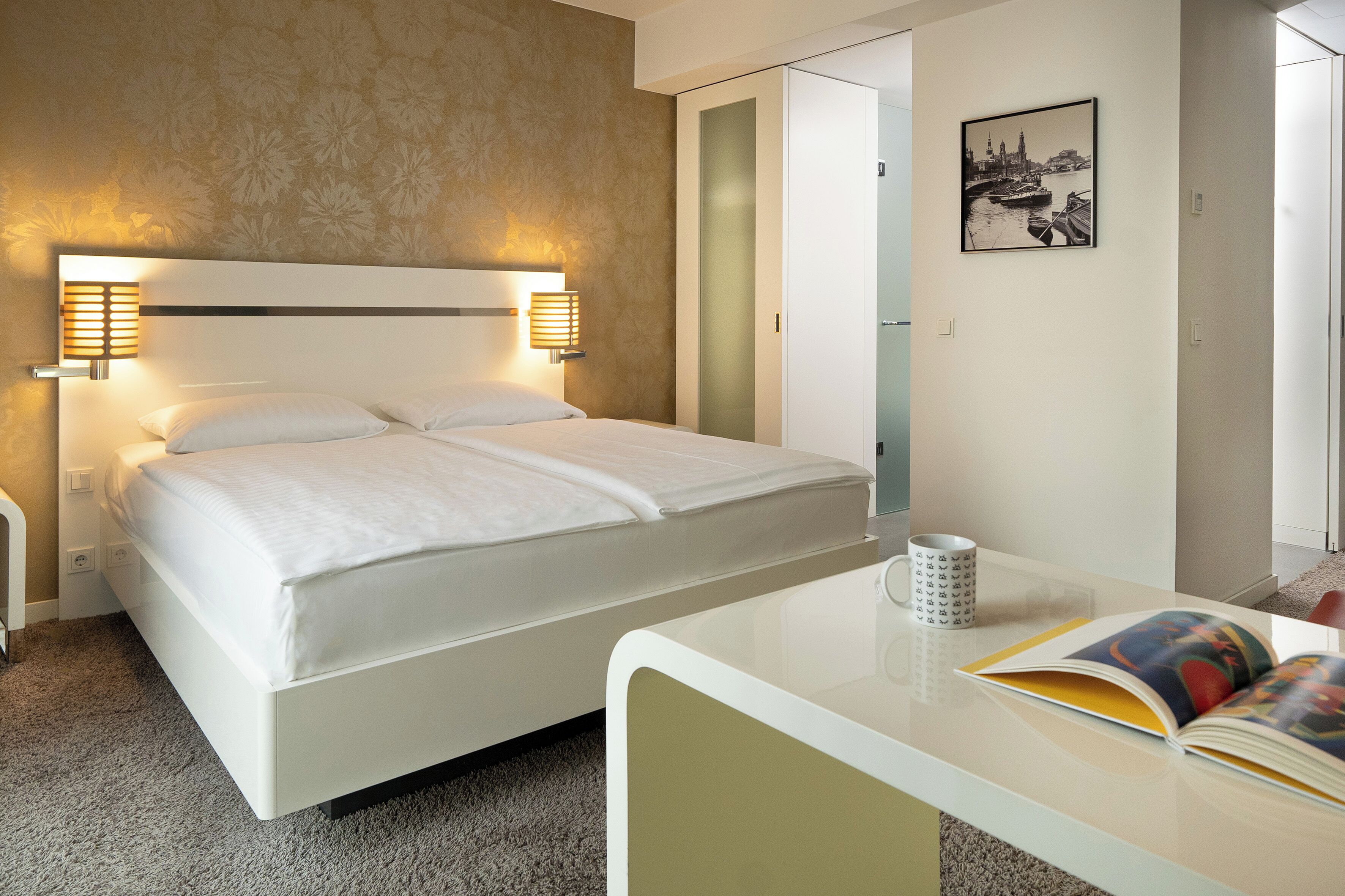 Une chambre d’hôtel avec un lit, des lampes de chevet, une table de chevet avec un livre, et un tableau encadré au mur.