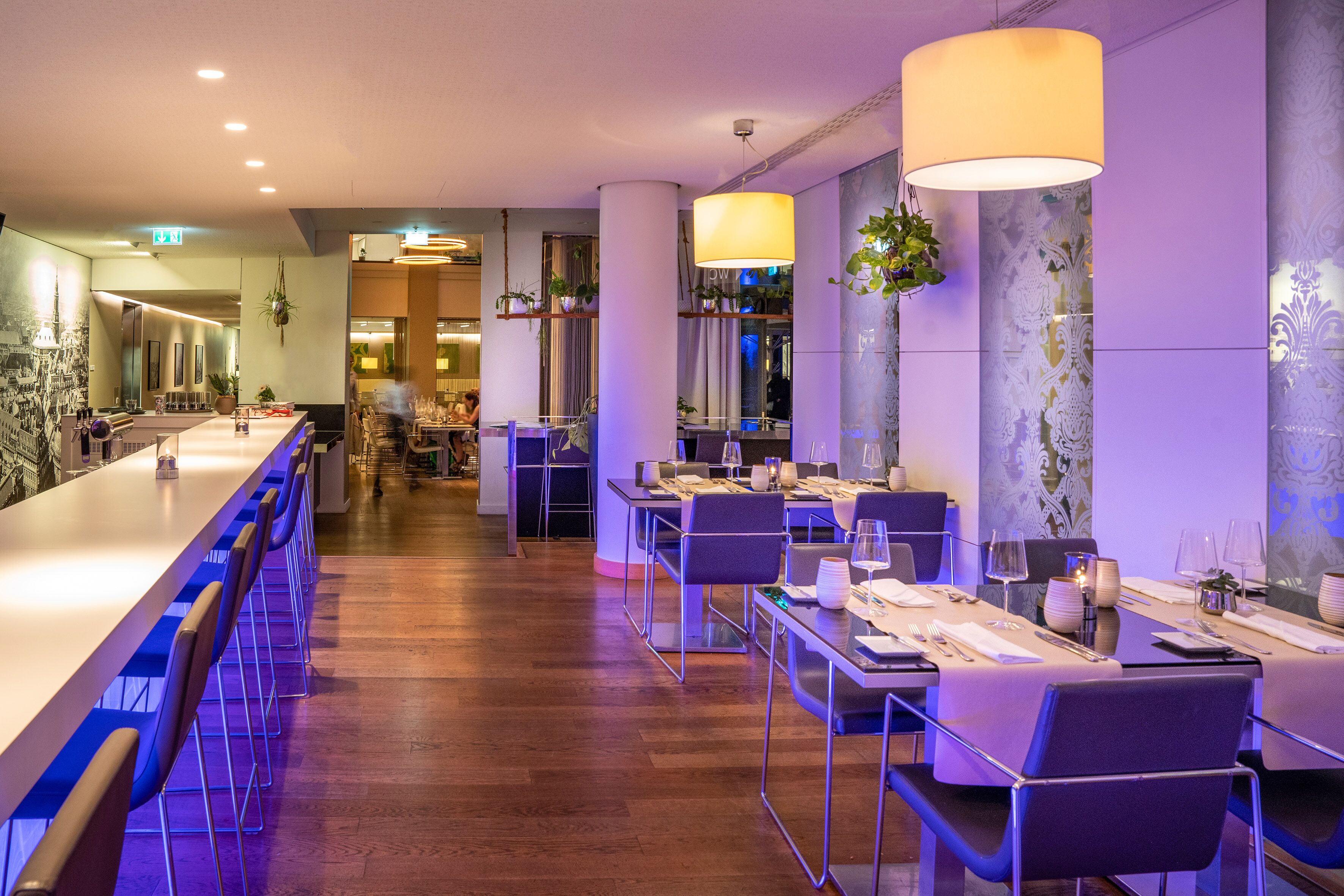 Ein modernes Restaurant mit langer Bar, mehreren Speisetischen und hängenden Pflanzen.