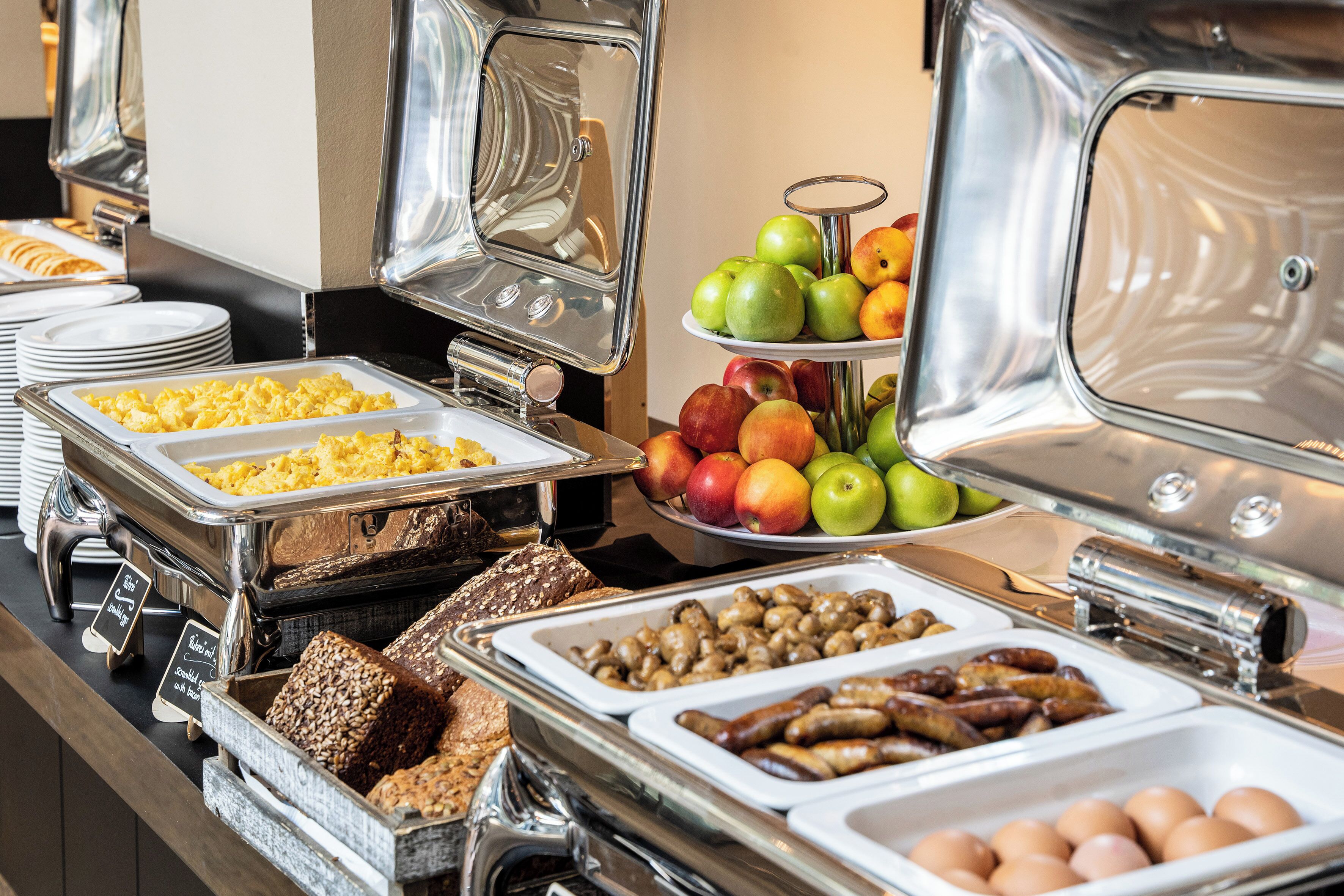 A buffet with various food items including fruits, eggs, sausages, and bread.