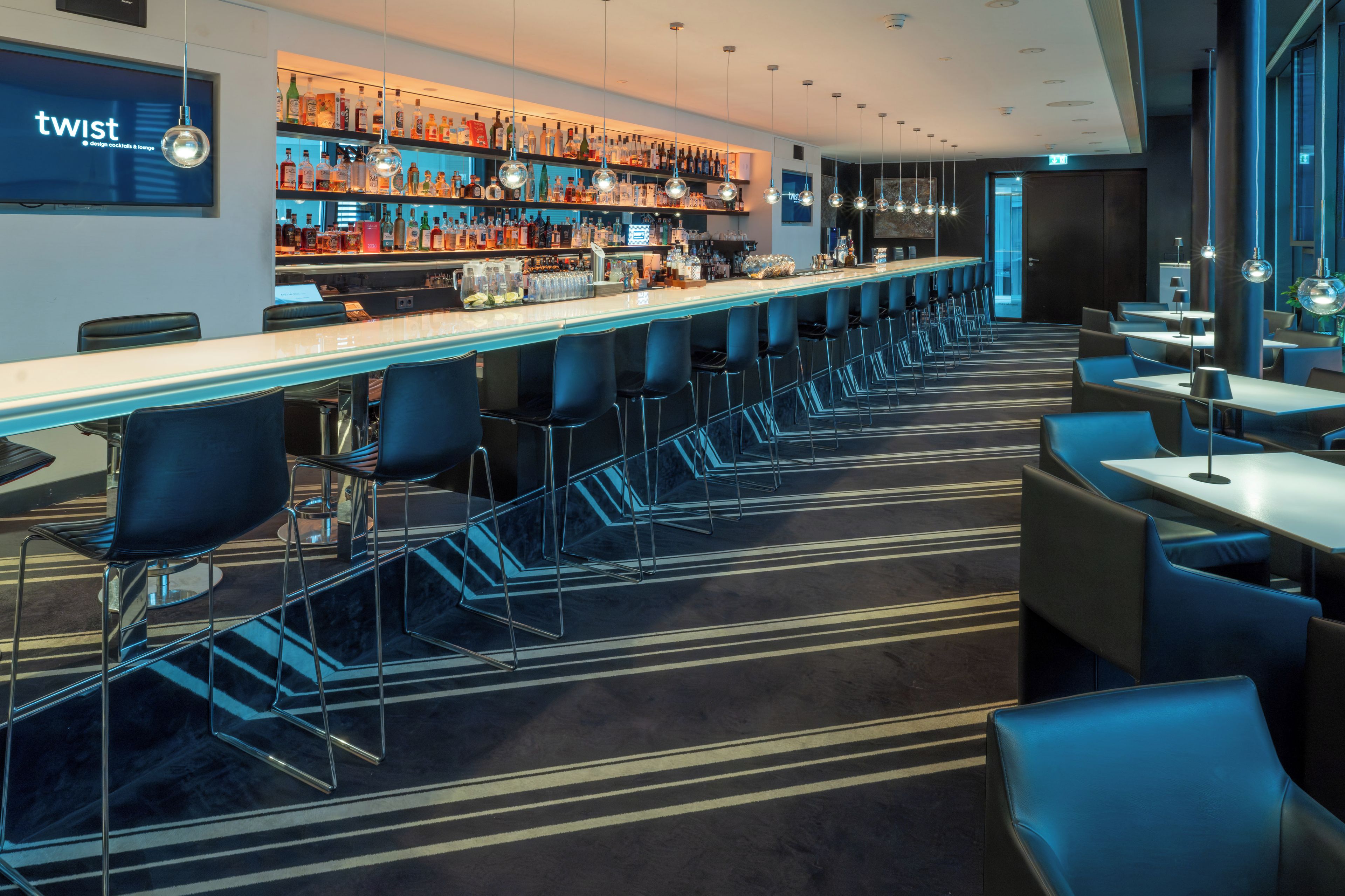 Uma área de bar com balcão extenso, vários bancos altos e variedade de garrafas e copos.