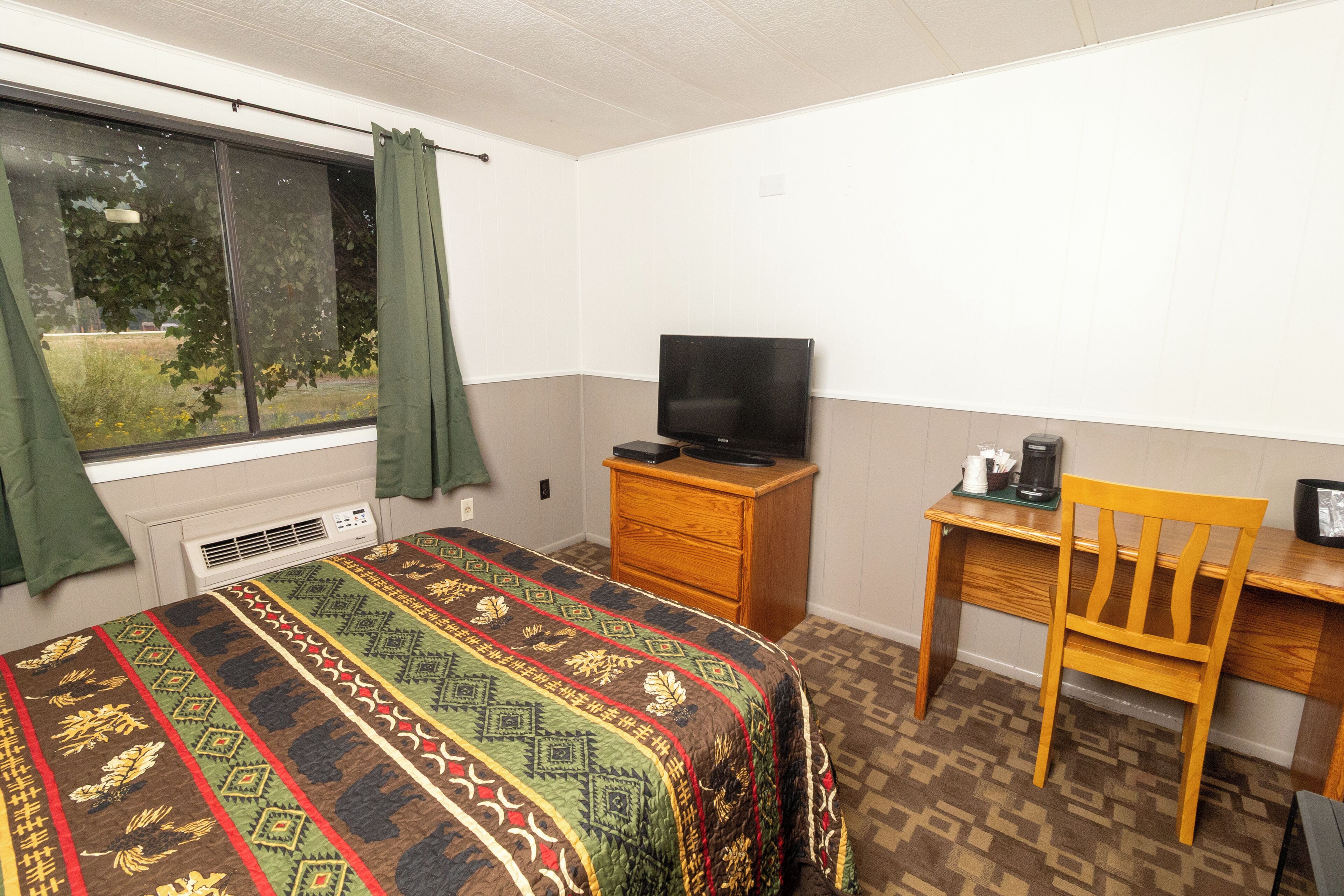 Un dormitorio con una cama, un televisor sobre un tocador de madera, un escritorio con una silla y una ventana con cortinas.