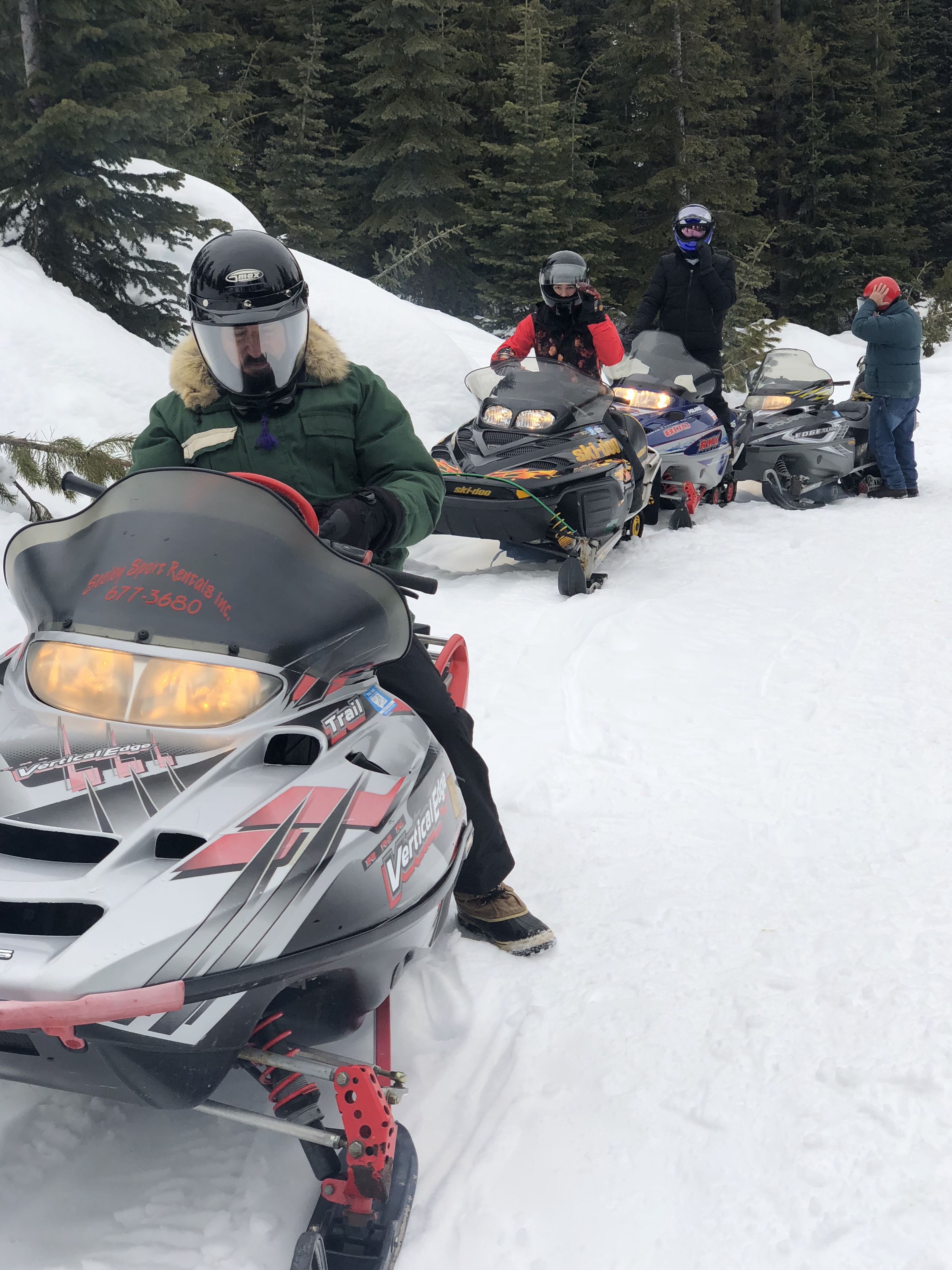 A group of snowmobiles parked in a snowy area with trees in the background.