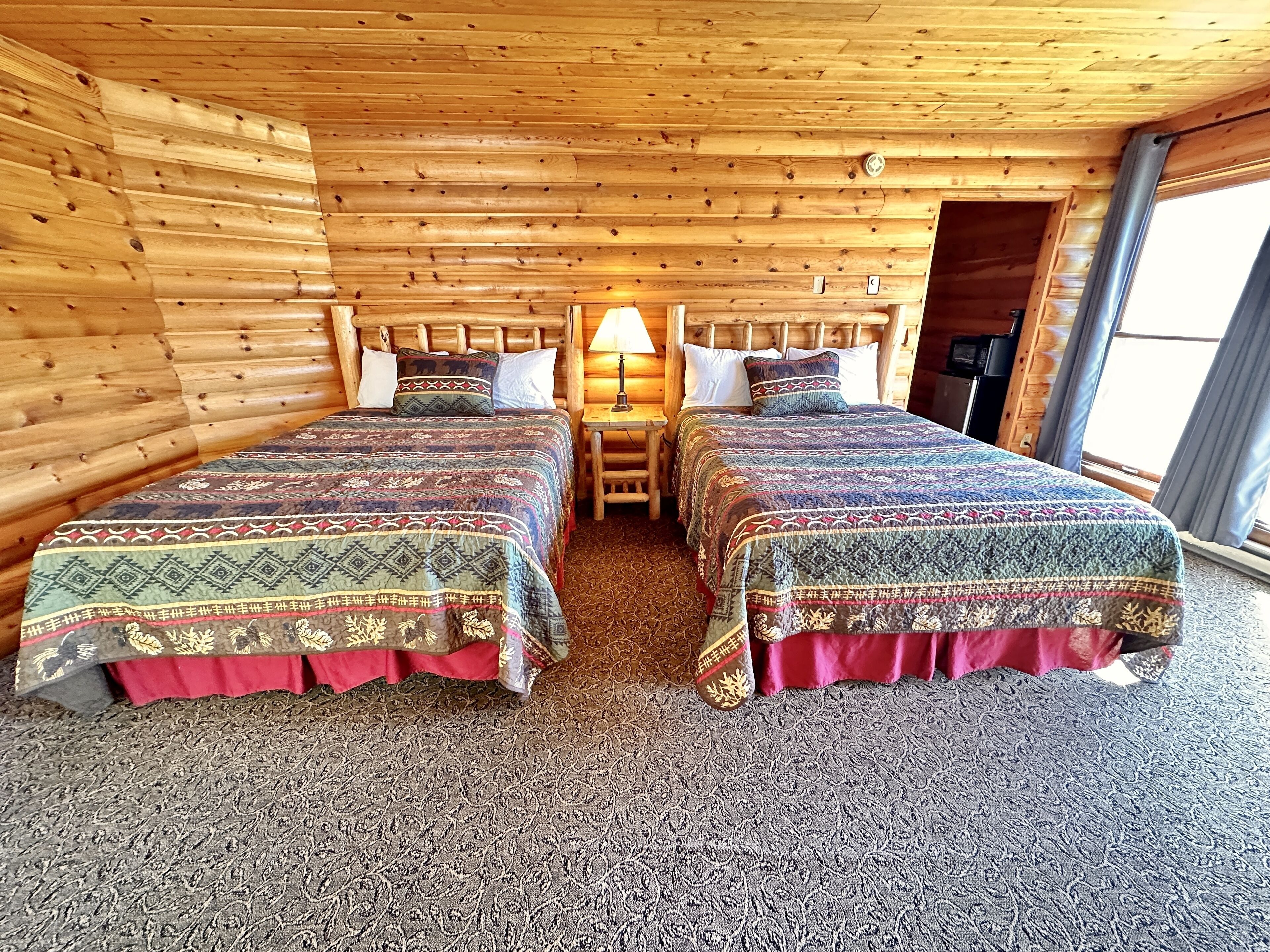 Una habitación de cabaña de madera con dos camas, una mesita de noche, una lámpara y una ventana.