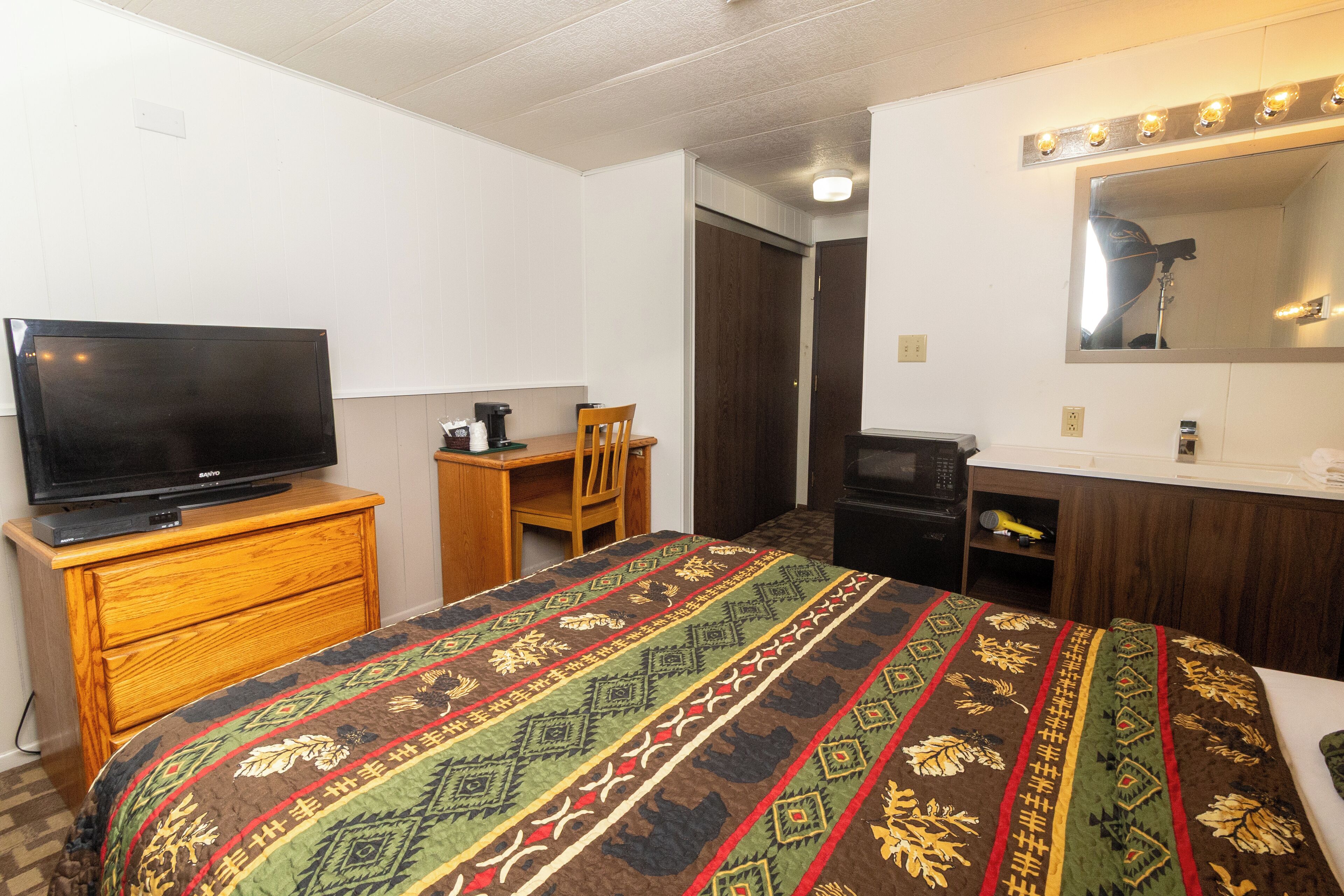 Een hotelkamer met een bed, een televisie op een houten dressoir, een bureau met een stoel, een spiegel met verlichting, en een deur naar een andere kamer.