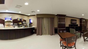 Un hall de hotel con una zona de desayuno autoservicio, un área de descanso con mesa y sillas, y un televisor montado en la pared.
