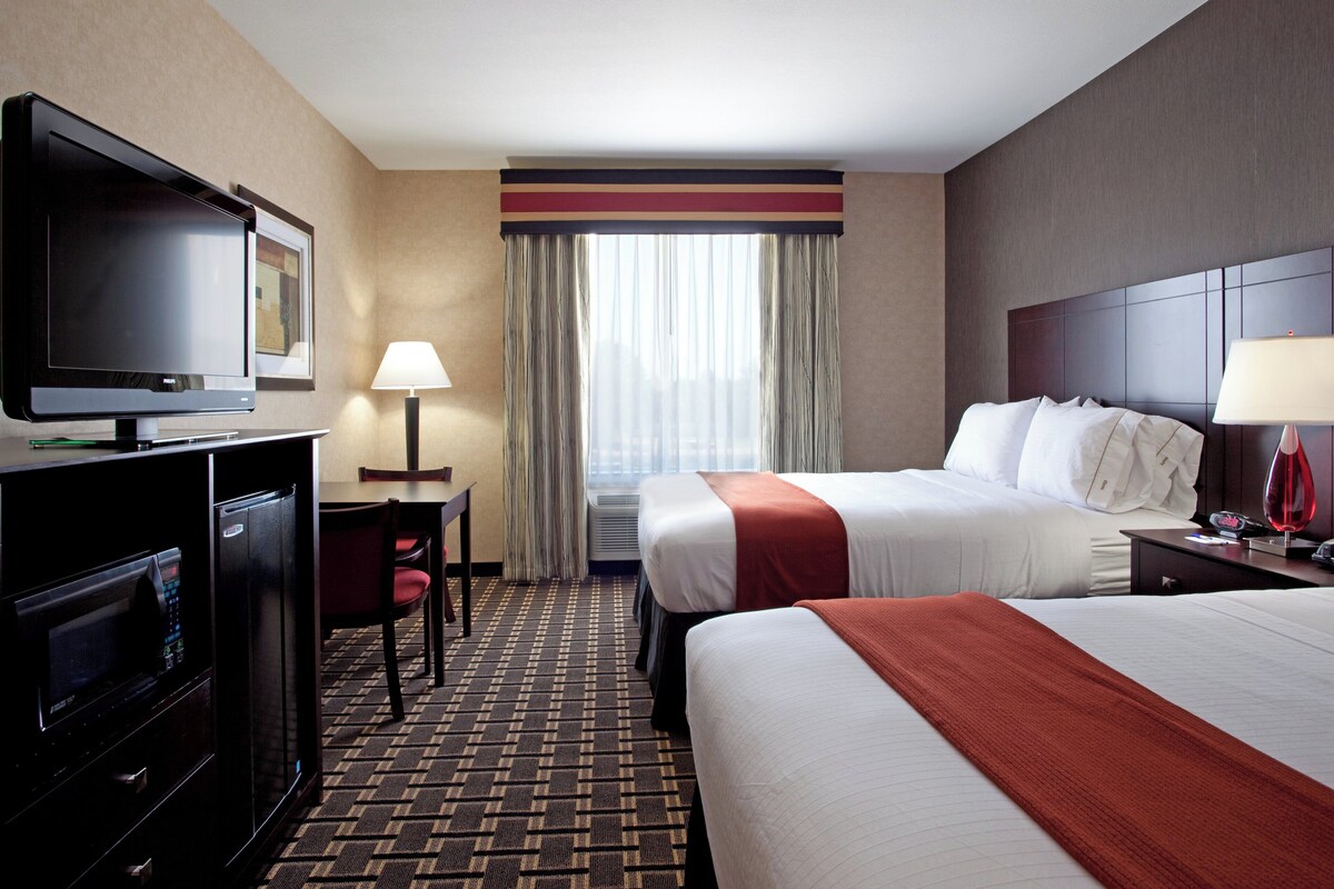 A hotel room with a TV, desk, chair, two beds, a lamp, and a window with curtains.