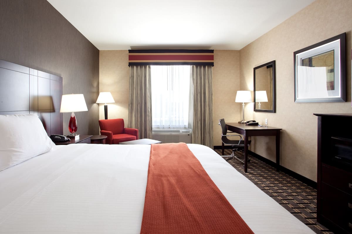 A hotel room with a large bed, two bedside lamps, a desk with a chair, a red chair, a large mirror, and a window with curtains.