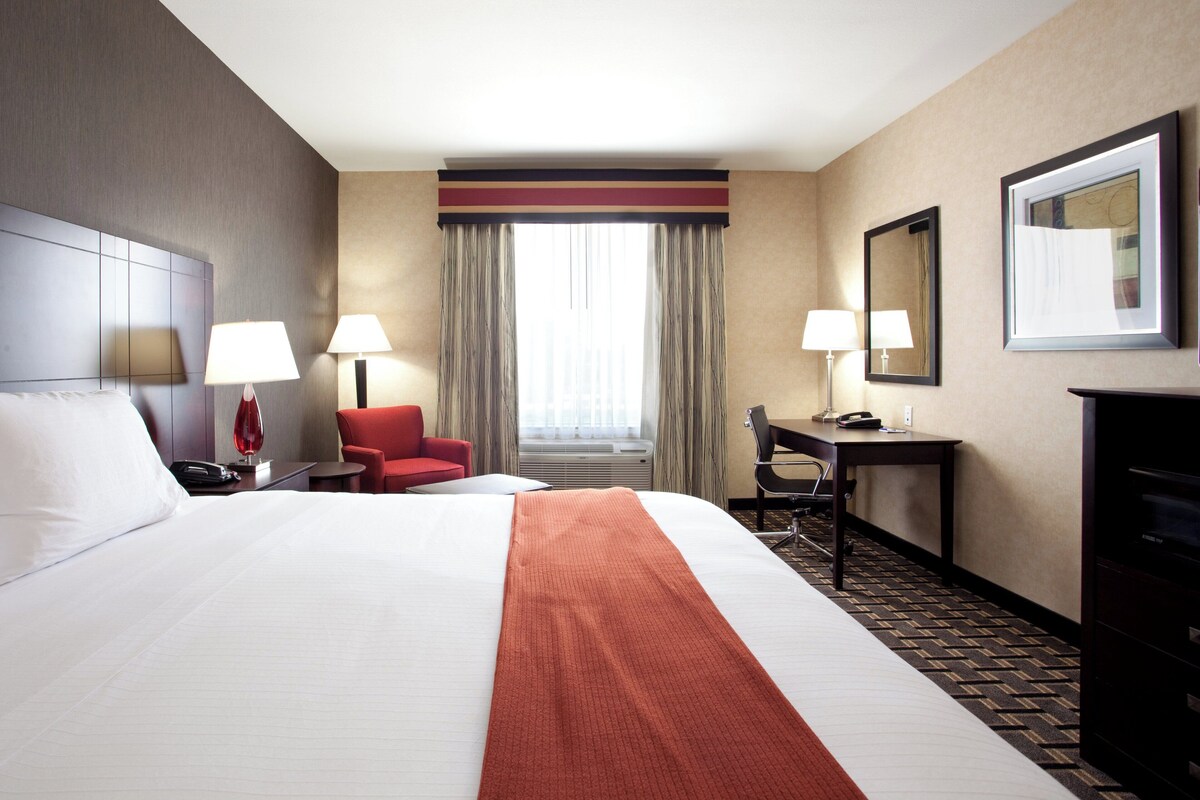 A hotel room with a large bed, two bedside lamps, a desk with a chair, a red armchair, a large mirror, and a window with curtains.