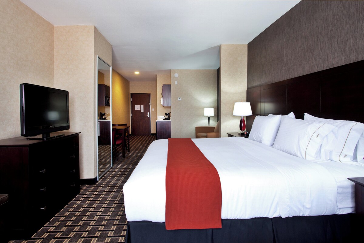 A hotel room with a large bed, a television on a dresser, a desk with a lamp, and a view into the kitchen area.