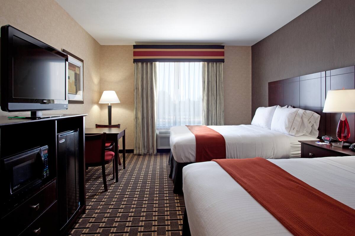 A hotel room with a television, desk, chair, two beds, and a window with curtains.