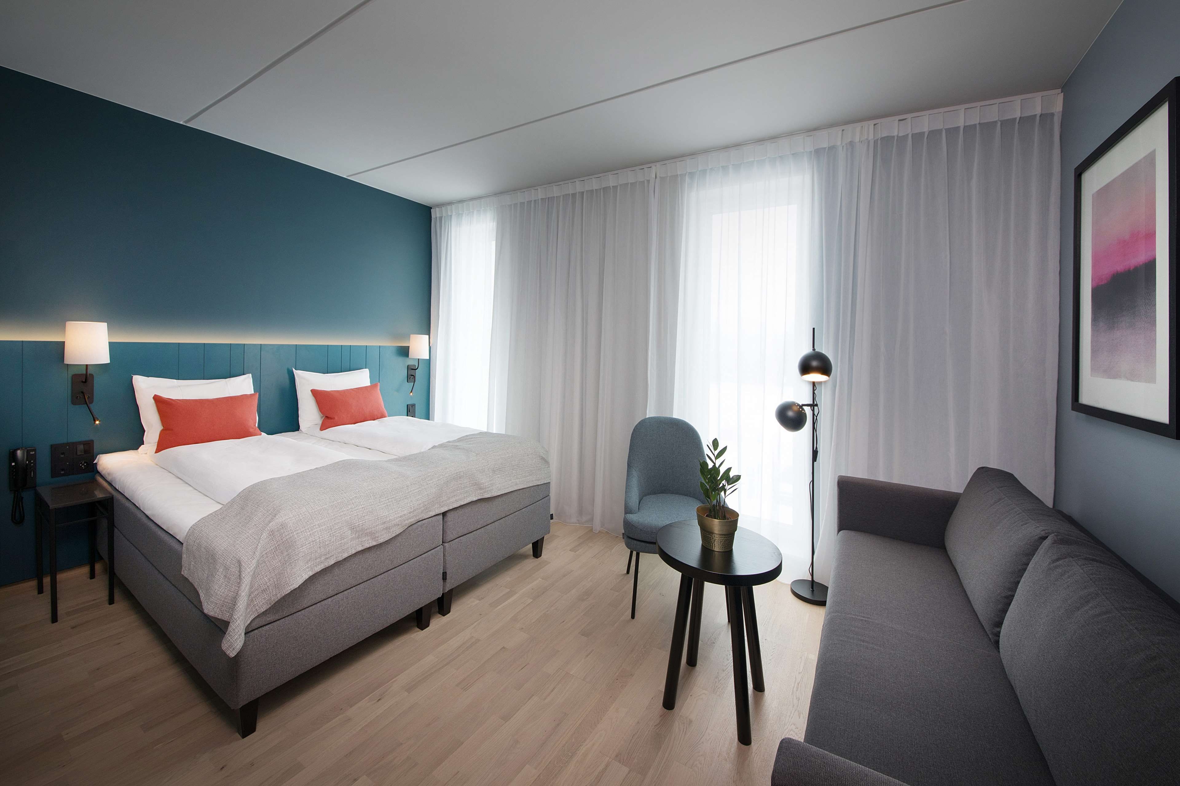 A modern hotel room with a bed, a sofa, a chair, a small table, and a floor lamp.