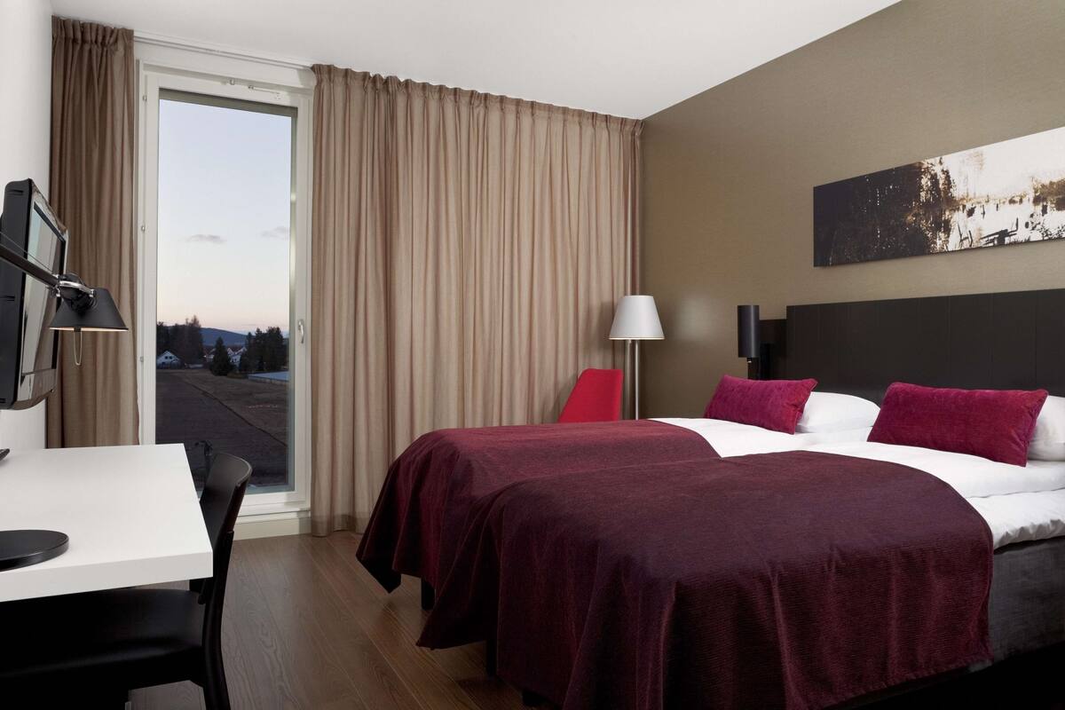 A hotel room with two beds, a desk, a chair, a lamp, and a view of the outside.