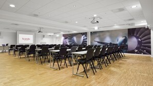 A conference room with rows of tables and chairs, a projector screen, and a large mural of a jet engine.
