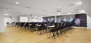 A conference room with rows of tables and chairs, a projector screen, and a large mural of a jet engine. - Scandic Oslo Airport (Nannestad)