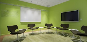 Ein Konferenzraum mit grünen Wänden, einer weißen Projektionsleinwand, einem Flachbildfernseher und modernen Stühlen, die um einen Tisch angeordnet sind.