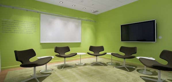 Un salón de conferencias con paredes verdes, una pantalla de proyección blanca, un televisor de pantalla plana y sillas modernas dispuestas alrededor de una mesa central.