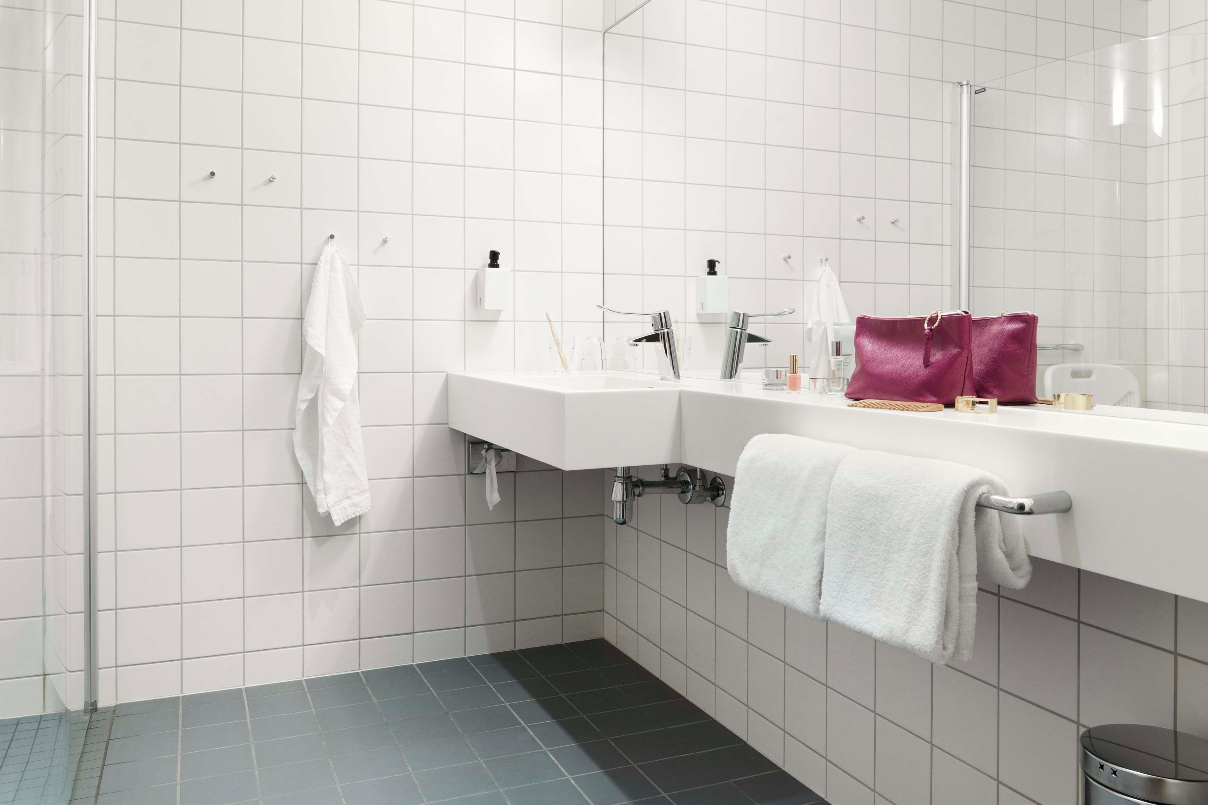 Un baño moderno con azulejos blancos, un espejo grande, un lavabo blanco y un bolso morado sobre el mostrador.