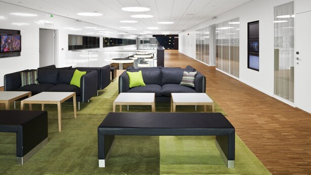 A modern office space with a long corridor, meeting tables, sofas, and a green carpet.