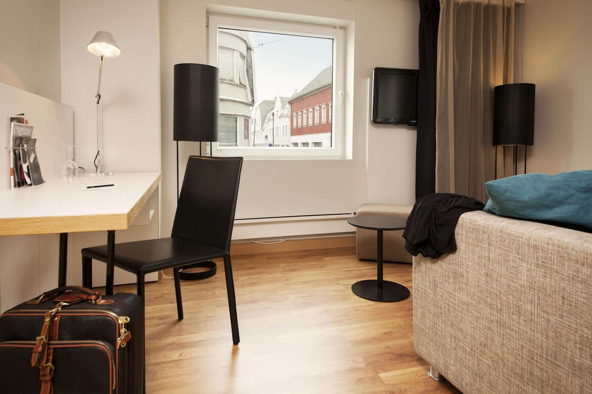 A hotel room with a desk, chair, and a bed. There is a window with a view of buildings outside.