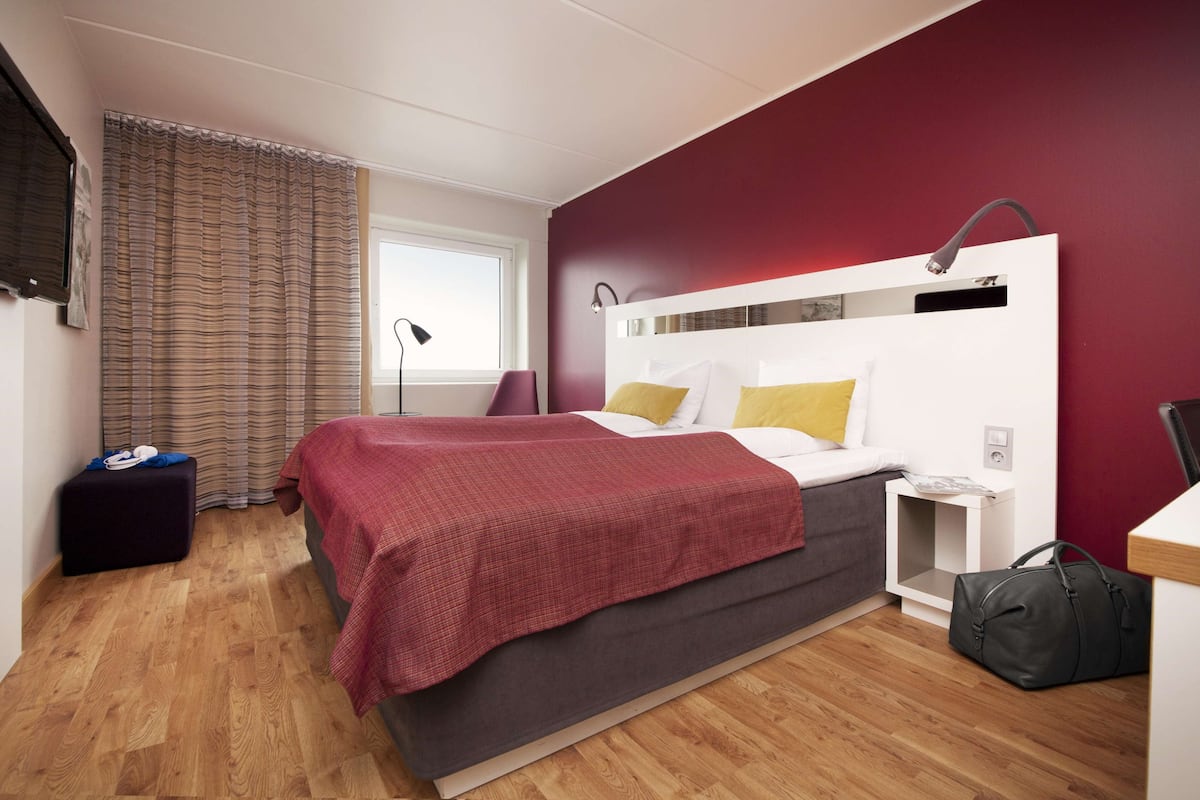 A modern hotel room with a large bed, a bedside table, a nightstand, a wardrobe, and a window with curtains.