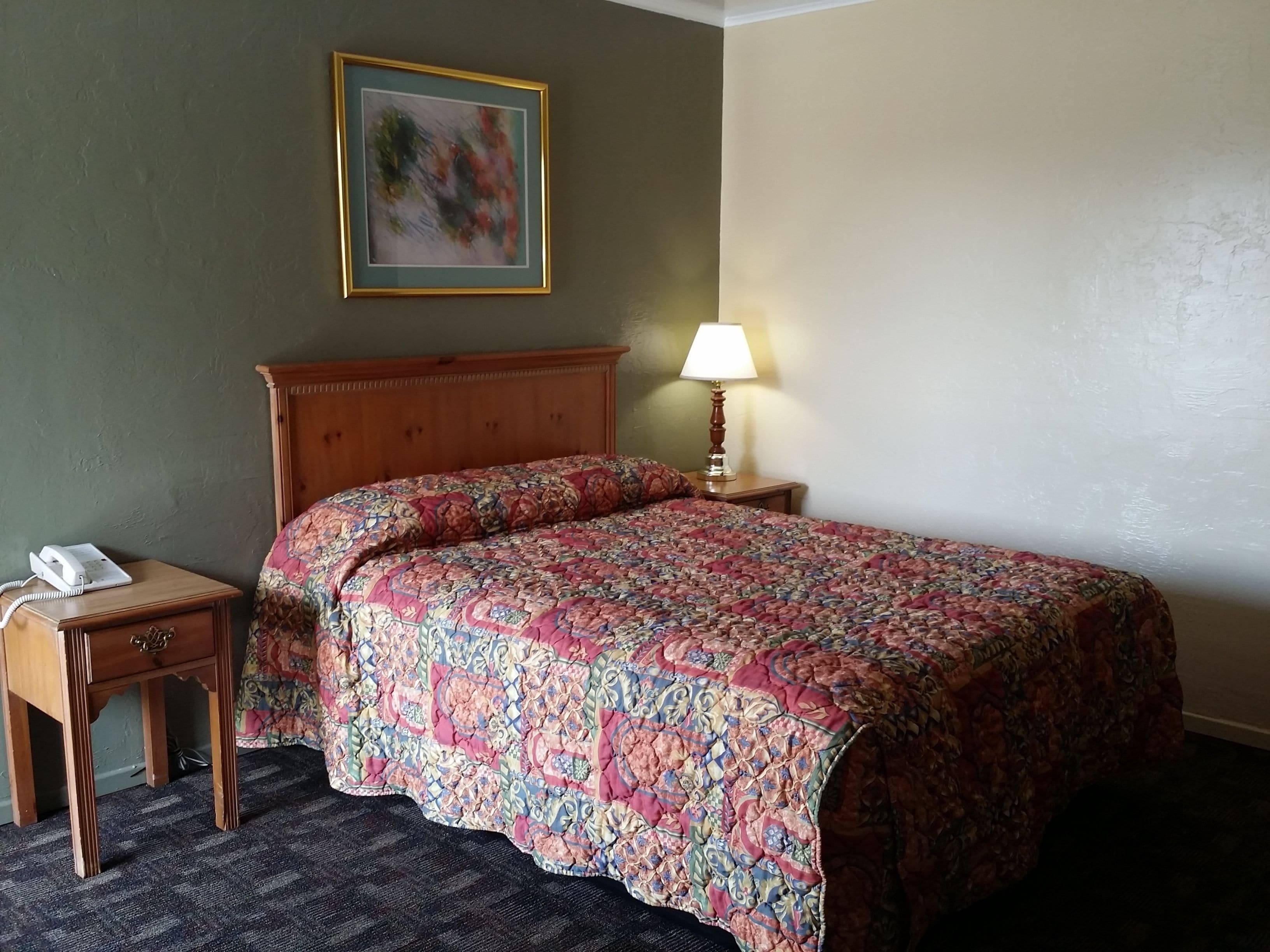 A hotel room with a wooden headboard, a floral-patterned bedspread, a bedside lamp, a telephone on a nightstand, and a framed picture on the wall.