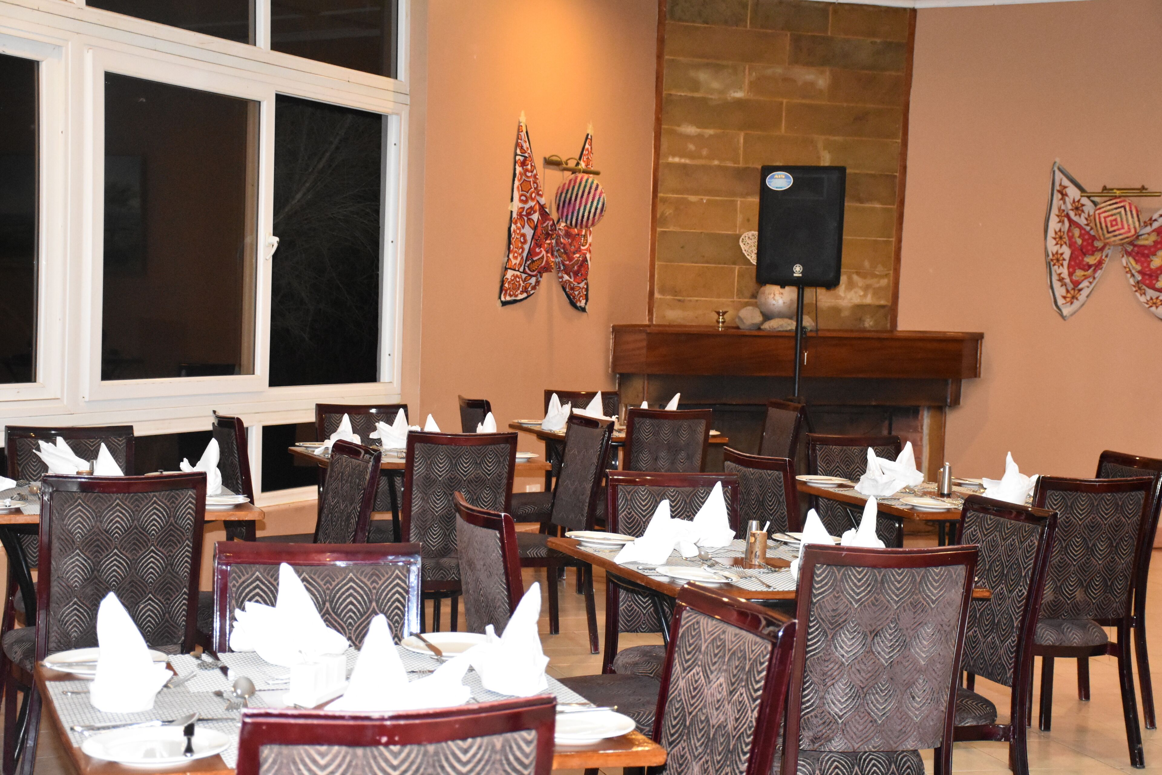 Uma sala de jantar com várias mesas preparadas para hóspedes, cadeiras com estofados estampados, uma lareira de tijolos e detalhes decorativos nas paredes.