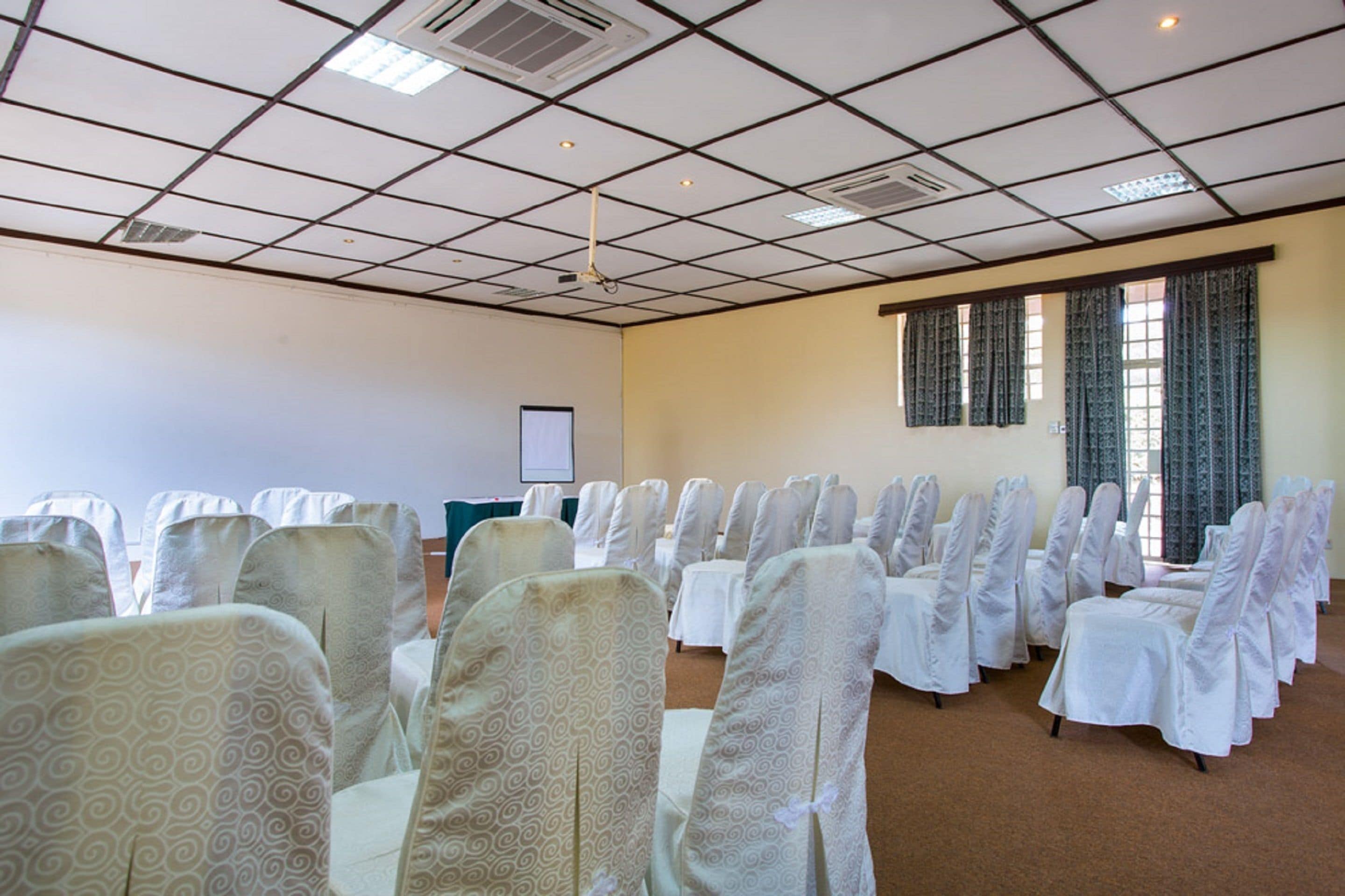 Un salón de conferencias con hileras de sillas cubiertas con manteles blancos y protectores blancos, una pantalla de proyector y un ventanal con cortinas.
