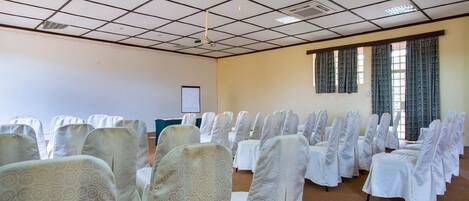 Un salón de conferencias con hileras de sillas cubiertas con manteles blancos y protectores blancos, una pantalla de proyector y un ventanal con cortinas.