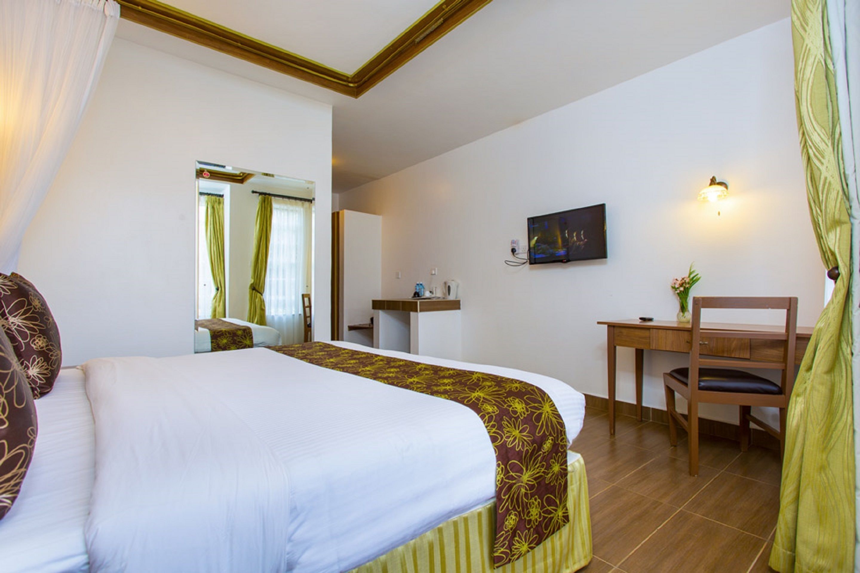 A hotel room with a large bed, a desk, a chair, a small table, a mirror, and a wall-mounted TV.
