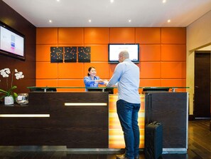 A hotel reception area with a reception desk, a digital screen displaying 'WELCOME,' and clocks on the wall.