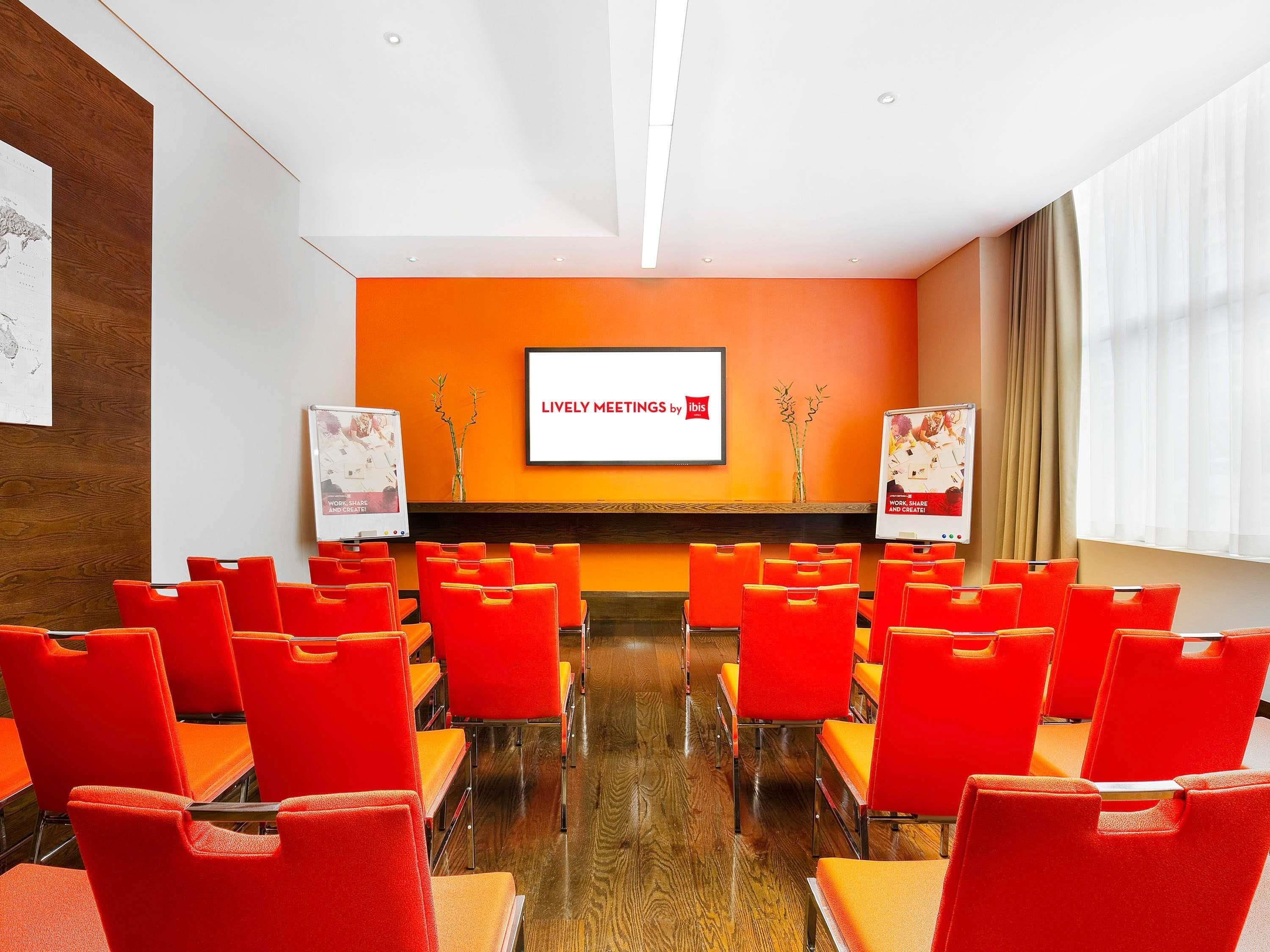 A conference room with orange chairs, a large screen displaying 'Lively Meetings by ibis,' and two posters on stands.