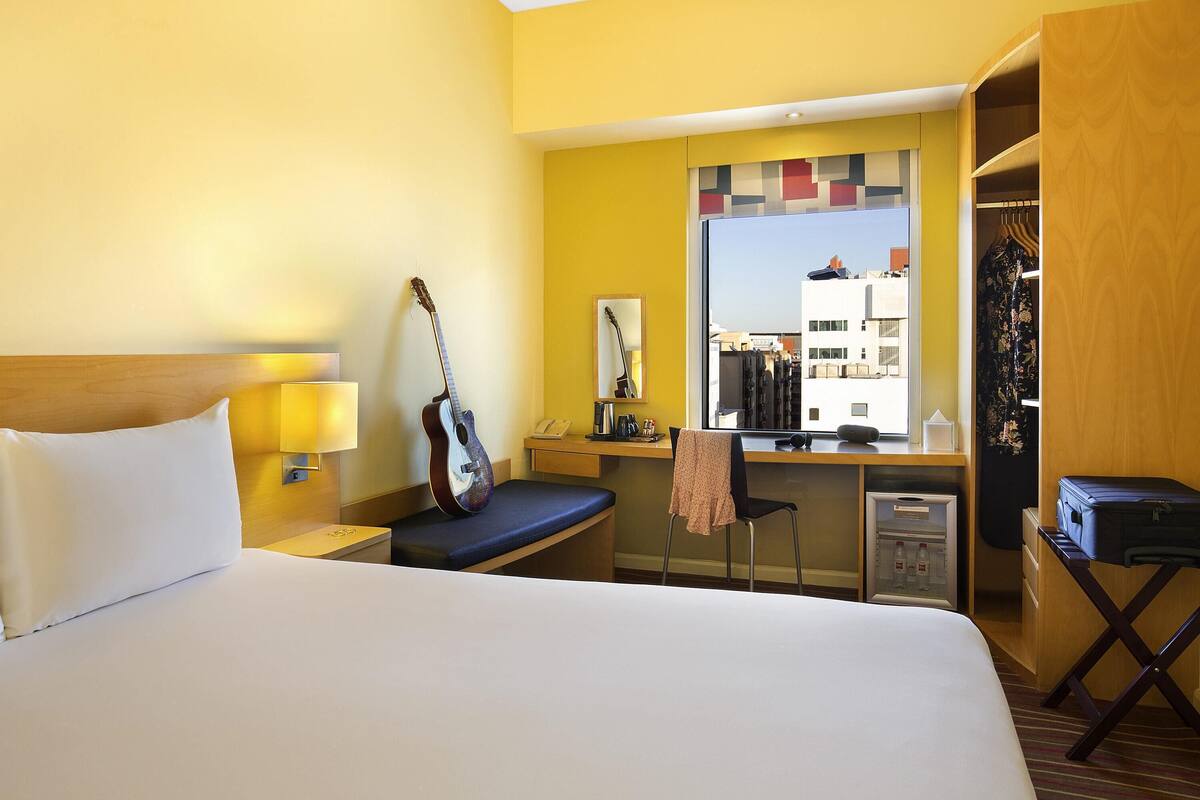 Une chambre d’hôtel avec un lit, un bureau, une guitare, un miroir et une vue sur la ville.