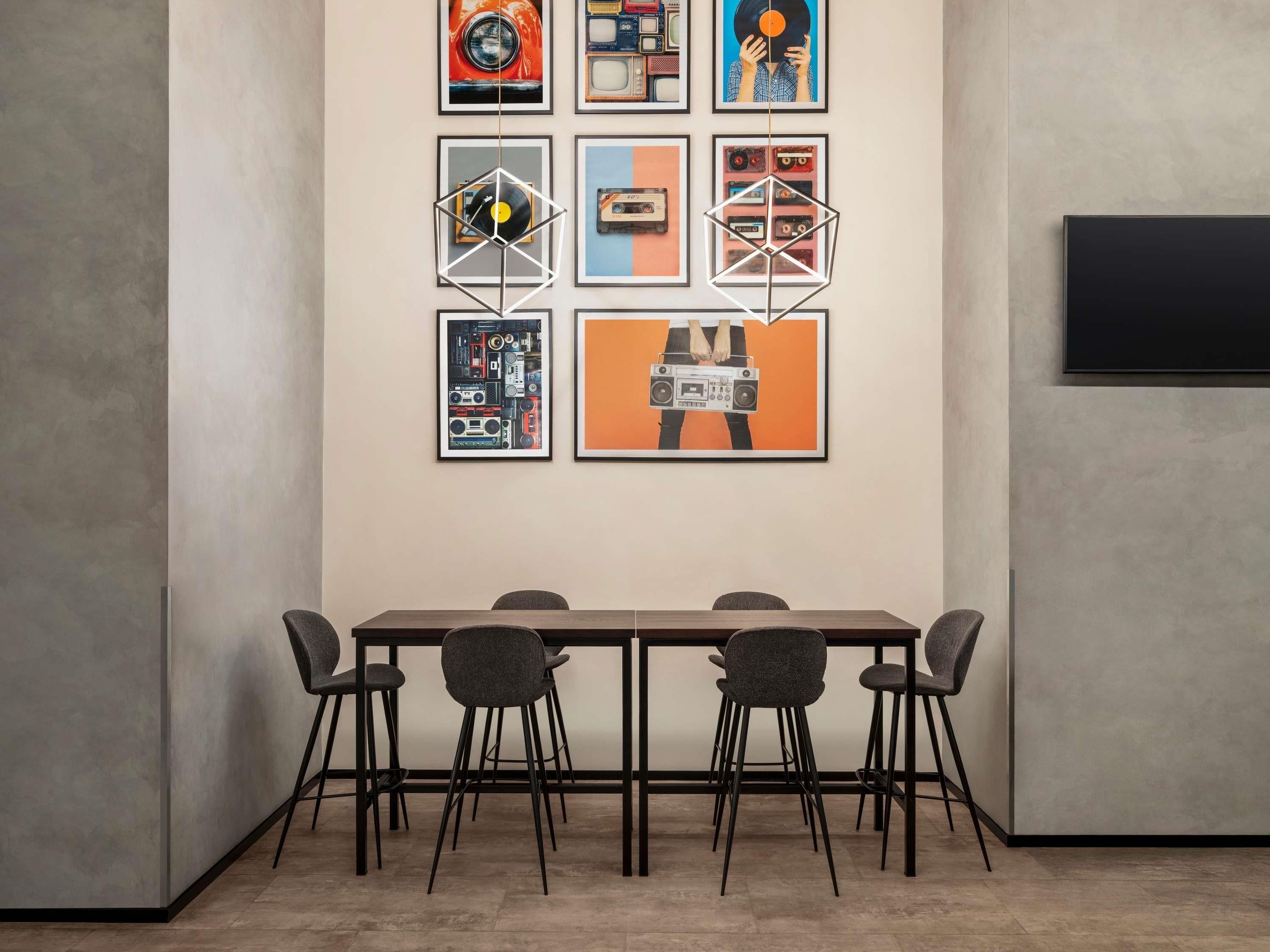A modern dining area with a long table and six chairs, framed artwork on the wall, and a flat-screen TV.