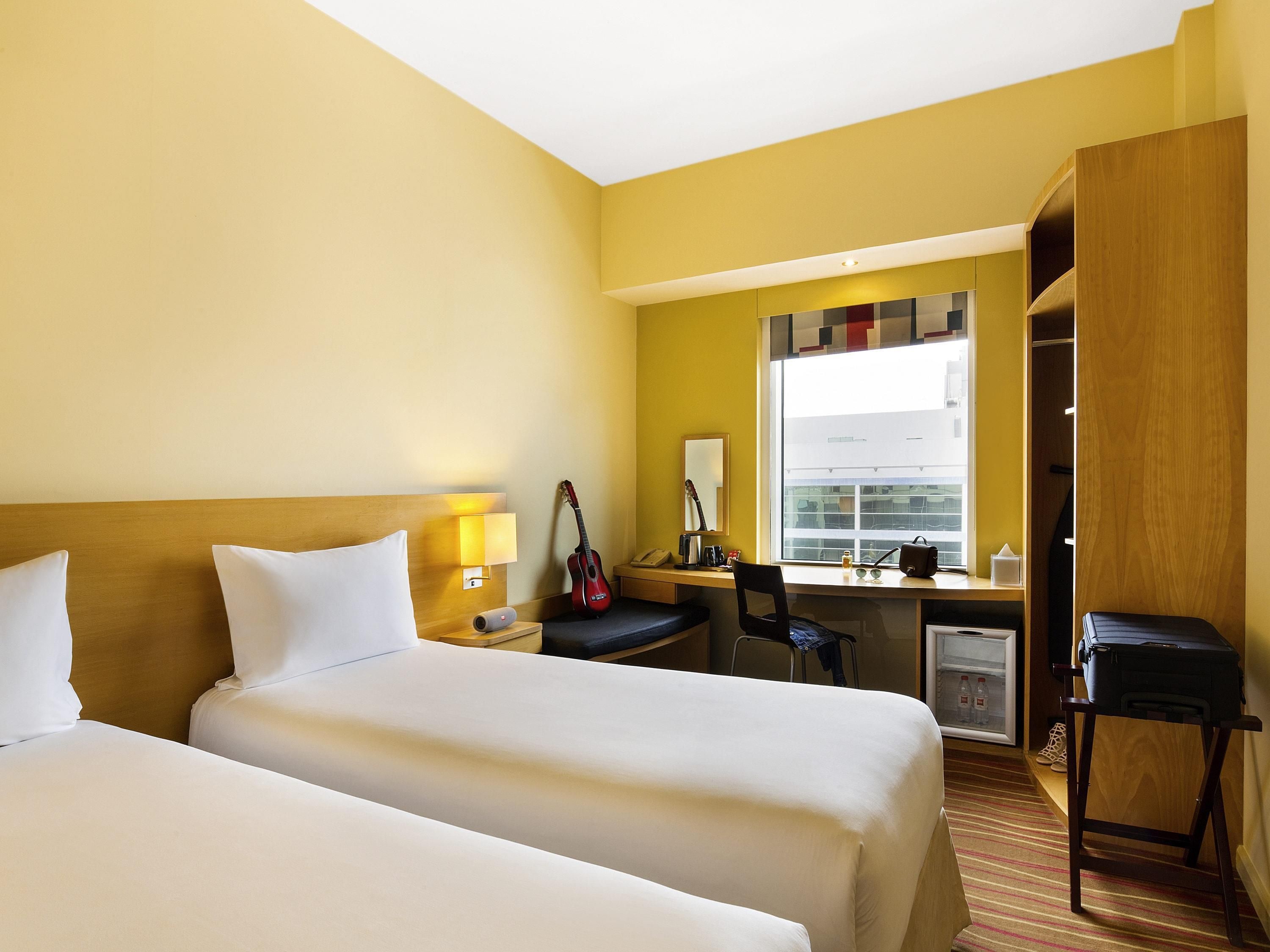 A hotel room with two beds, a desk, a chair, a wardrobe, and a window with a view of buildings.